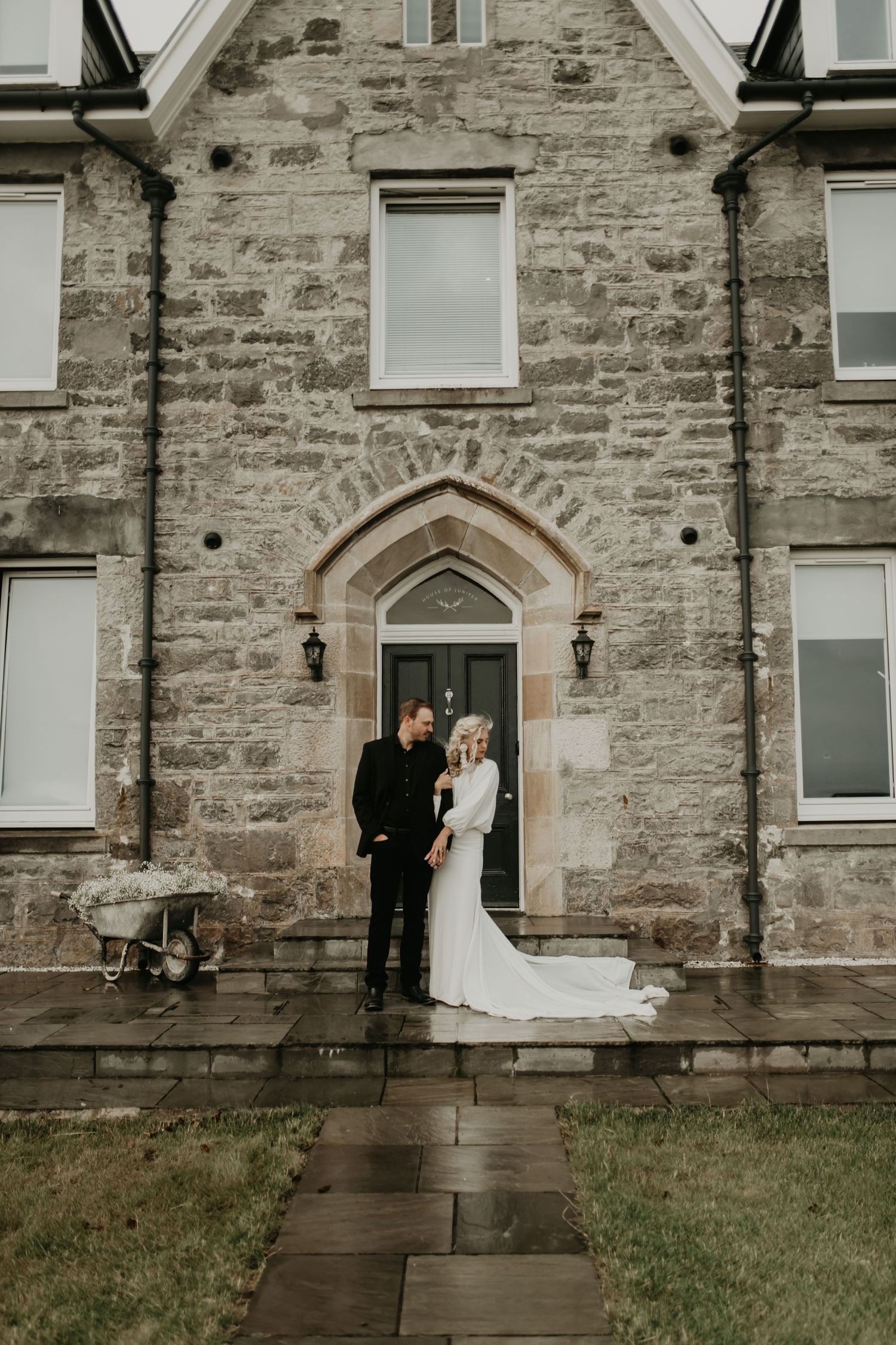 A couple stands in formal wedding attire in front of a stone building featuring an arched Gothic doorway entrance in isle of skye Scotland.
