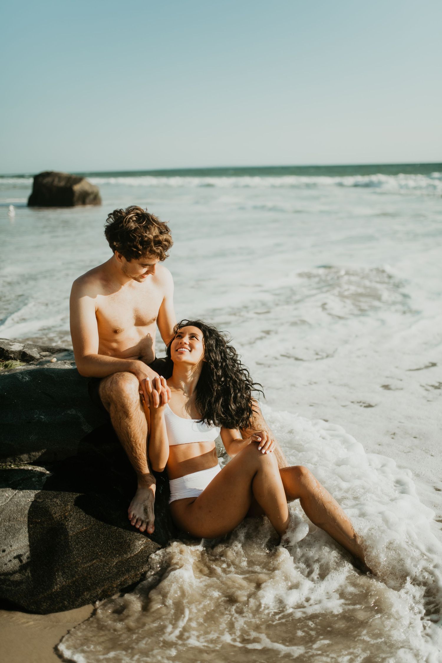 A couple in swimwear sits intimately on coastal rocks as ocean waves crash around them at the beach.