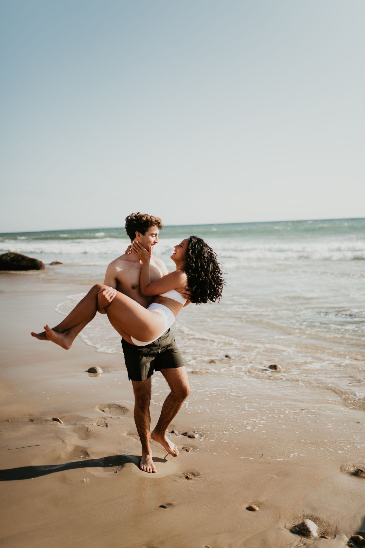 A romantic beach scene with a couple in swimwear embracing playfully on the sand near the ocean.