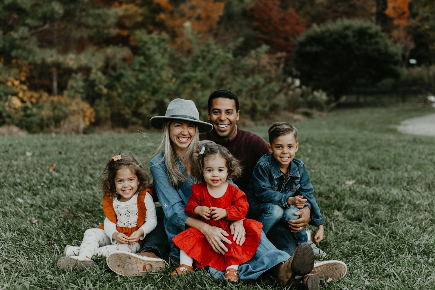 A family sits together on grassy ground during autumn with fall foliage visible in the background.