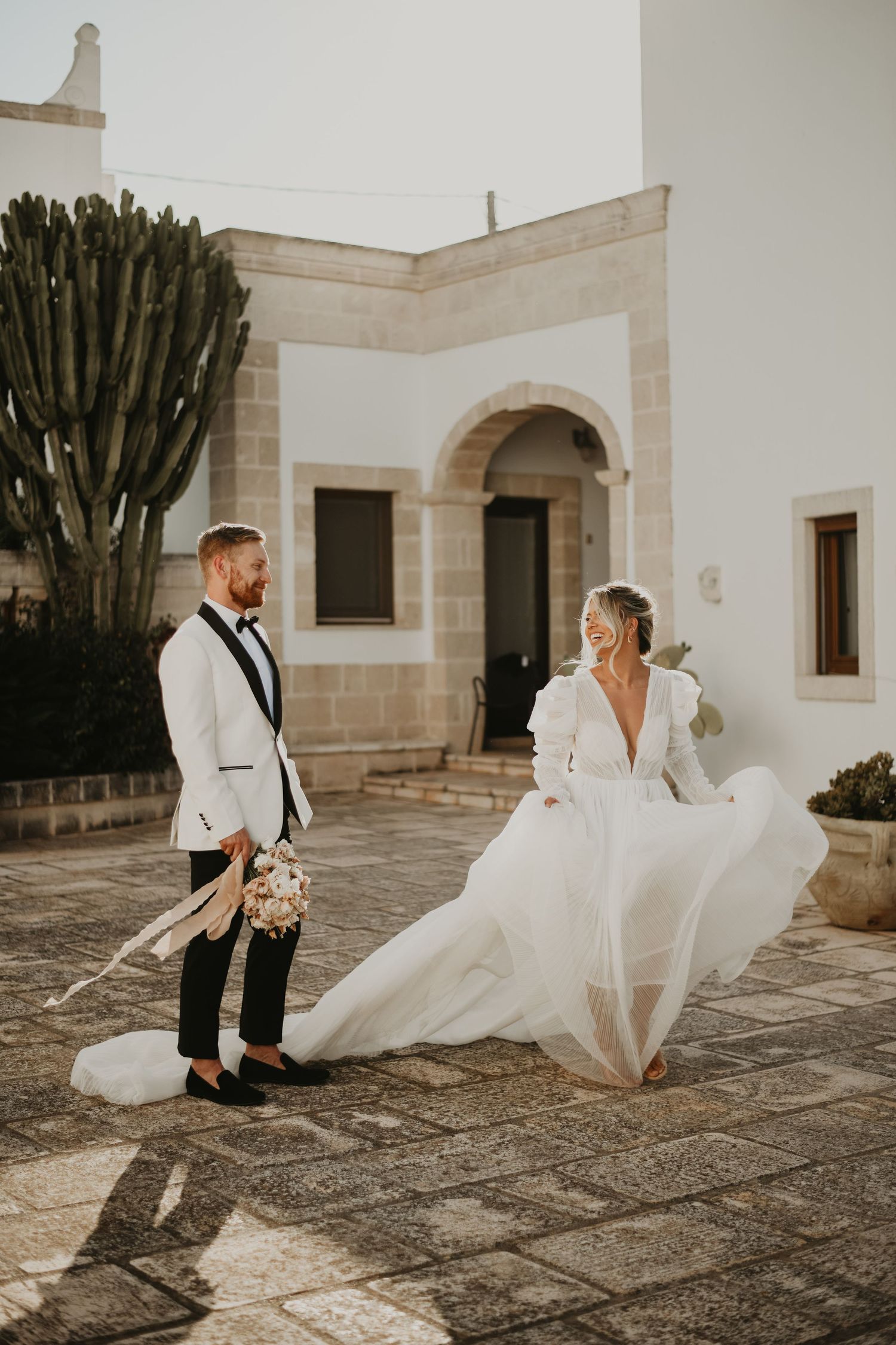 A couple in elegant wedding attire poses in front of a Mediterranean-style building in Italy.