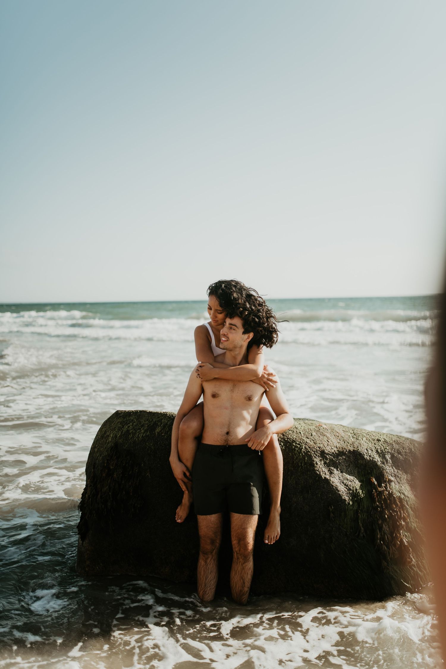 Parent and child share a tender moment while standing on rocks at the beach as waves crash around them.