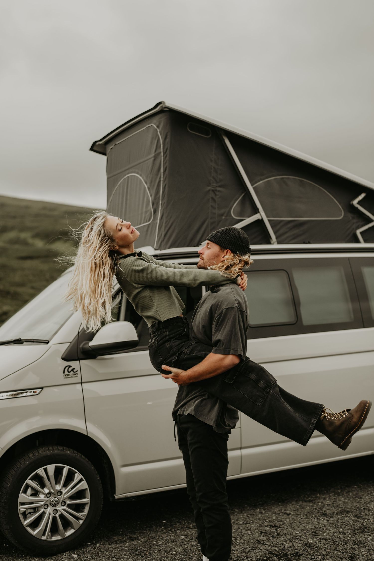 Couple enjoying adventure in front of their silver camper van with pop-up roof against moody overcast sky.