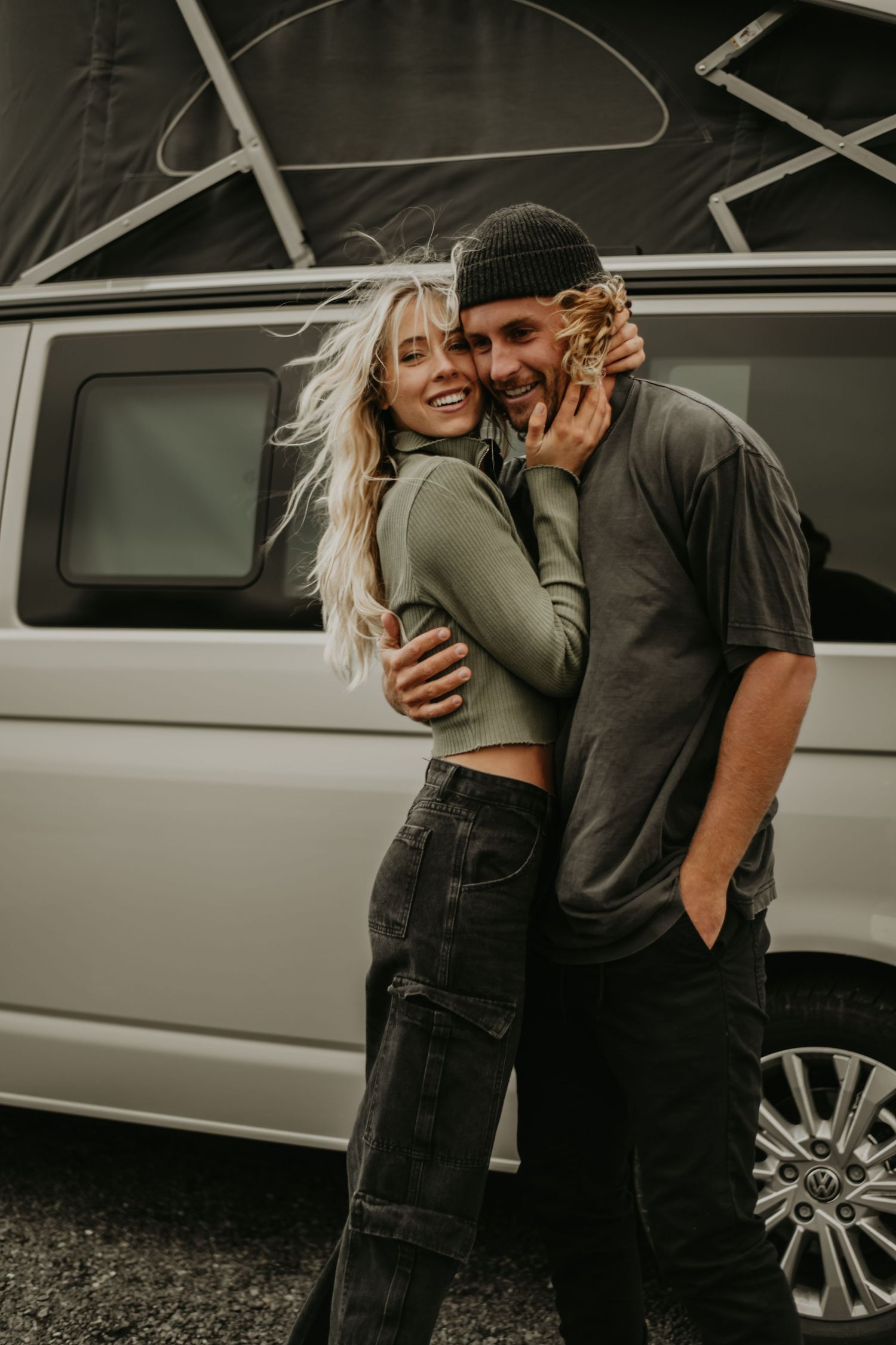 Couple embraces lovingly in front of silver van on overcast day wearing casual green and black outfits.