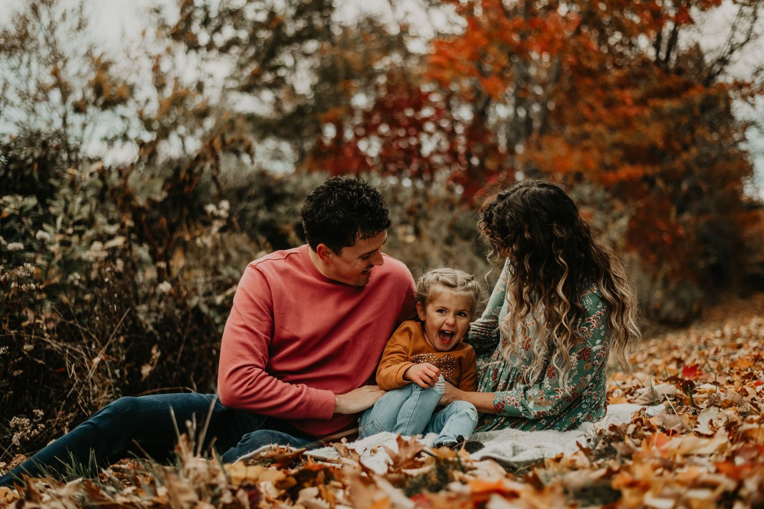 A family spends quality time together on a blanket surrounded by vibrant autumn leaves in a peaceful outdoor setting.