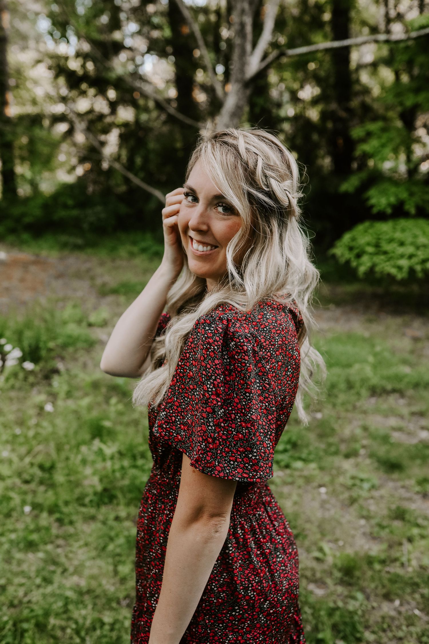 A woman in a red floral dress smiles while touching her face in a sunlit forest setting.