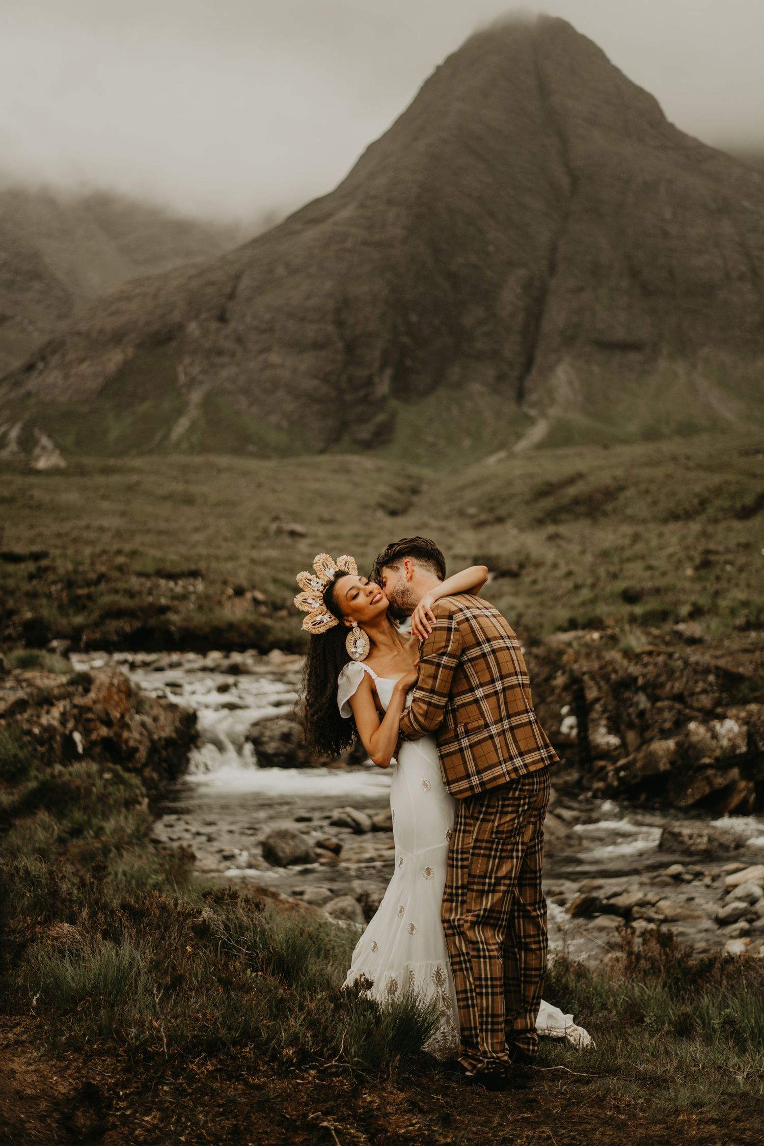 Couple shares romantic moments by scenic mountain stream in remote wilderness landscape during wedding photoshoot in Isle of Skye Scotland.