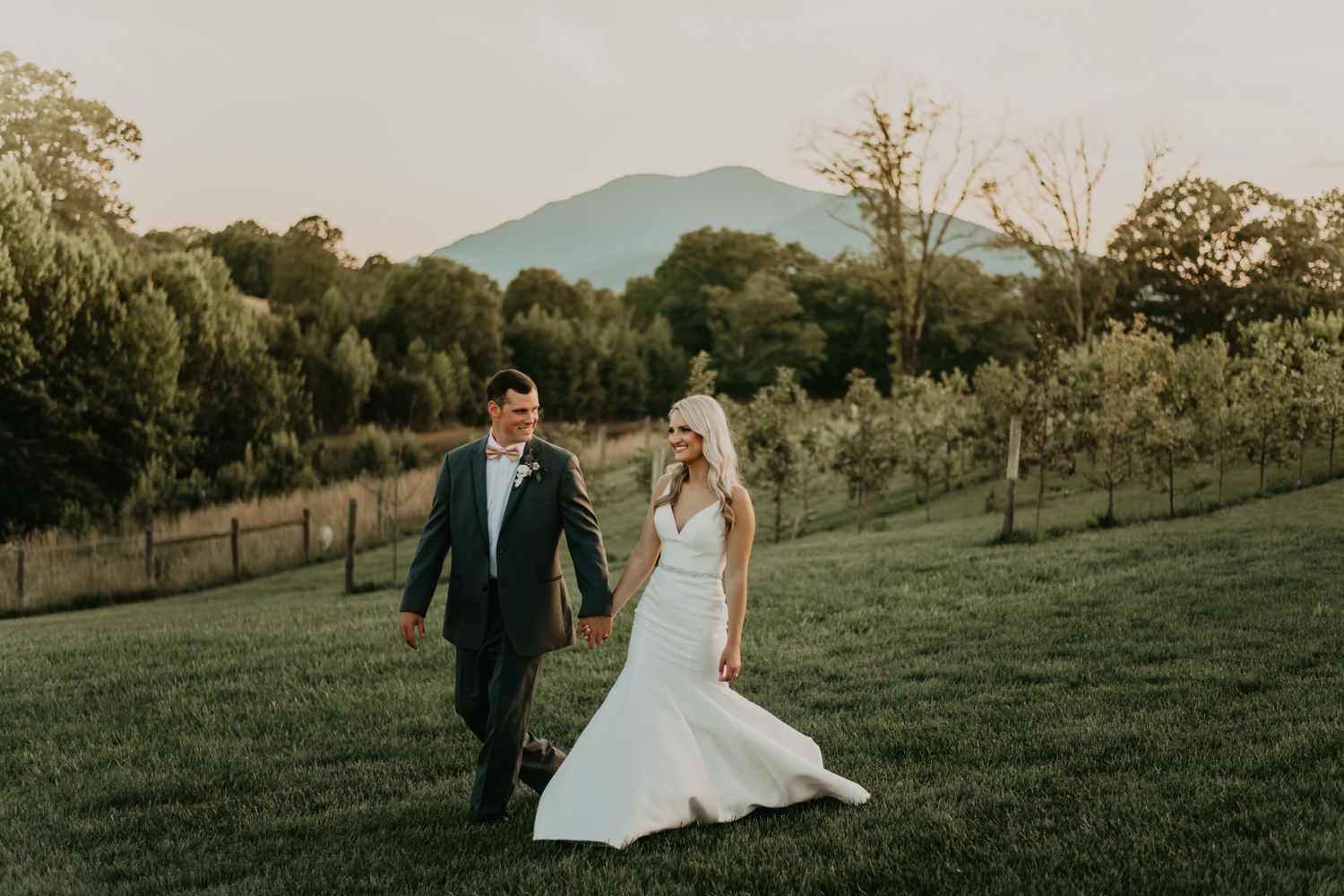 Newlyweds walk through rolling green hills with mountain backdrop at sunset in North Carolina.