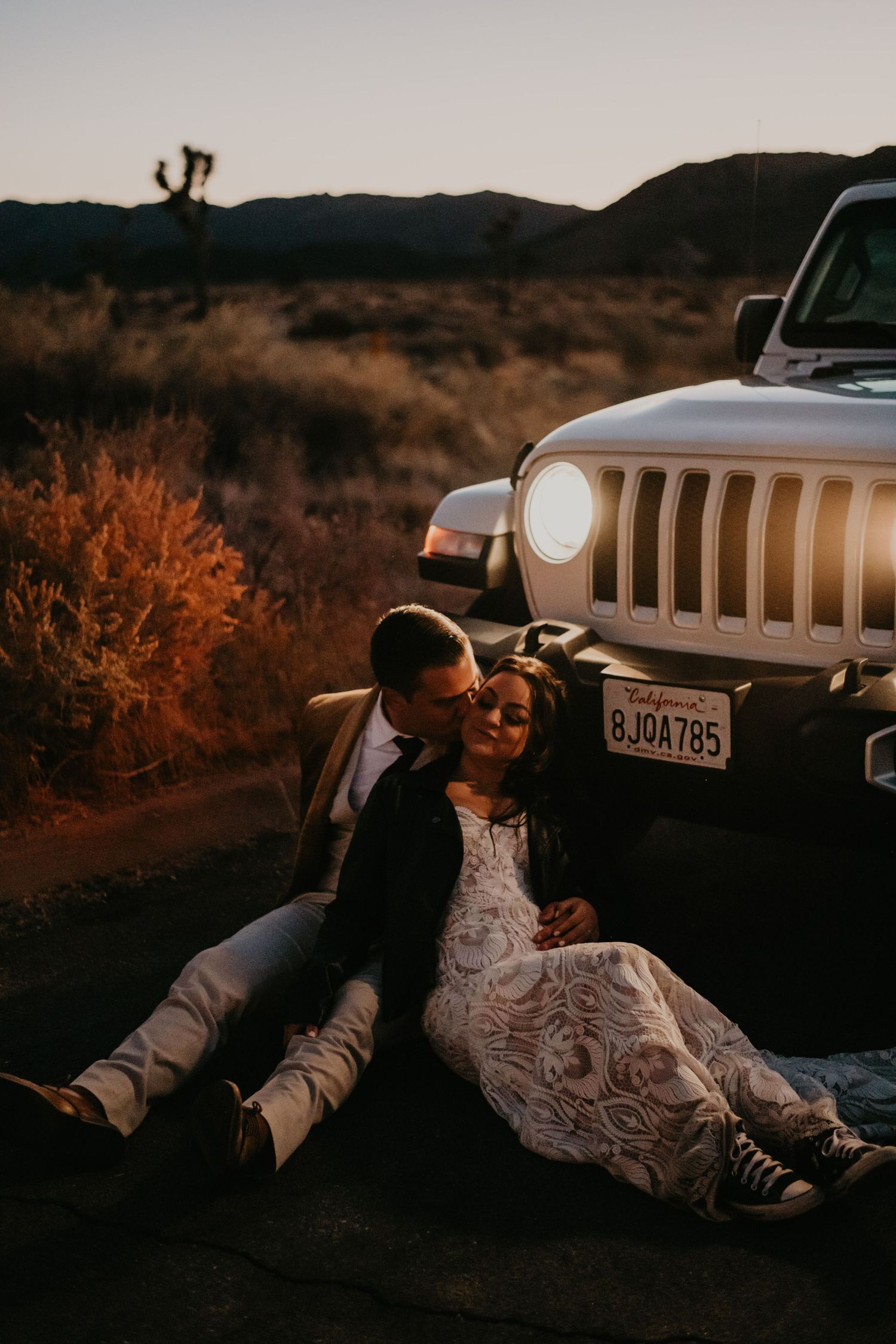 A couple in wedding attire sits against a Jeep at sunset in a desert landscape in Joshua tree, California.