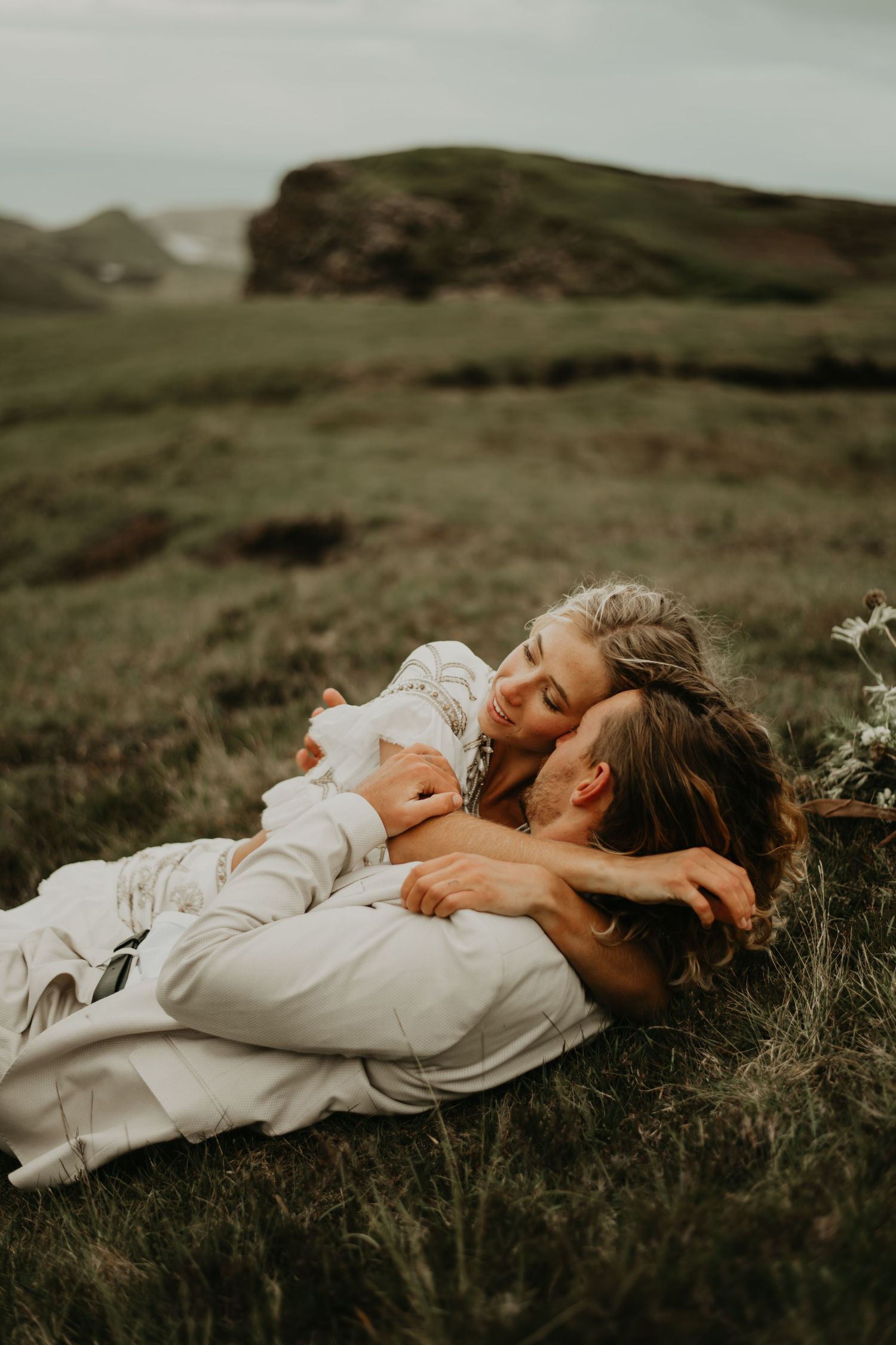 A romantic couple in white clothing lying together on a grassy hillside at sunset in Isle of Skye Scotland.