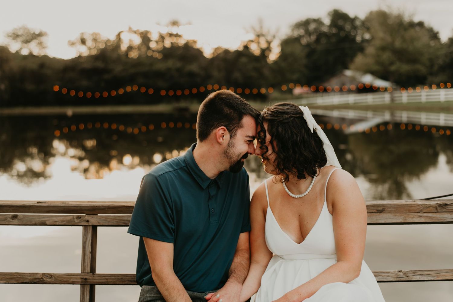A romantic couple shares an intimate moment at sunset by a lake with string lights creating a warm ambiance.