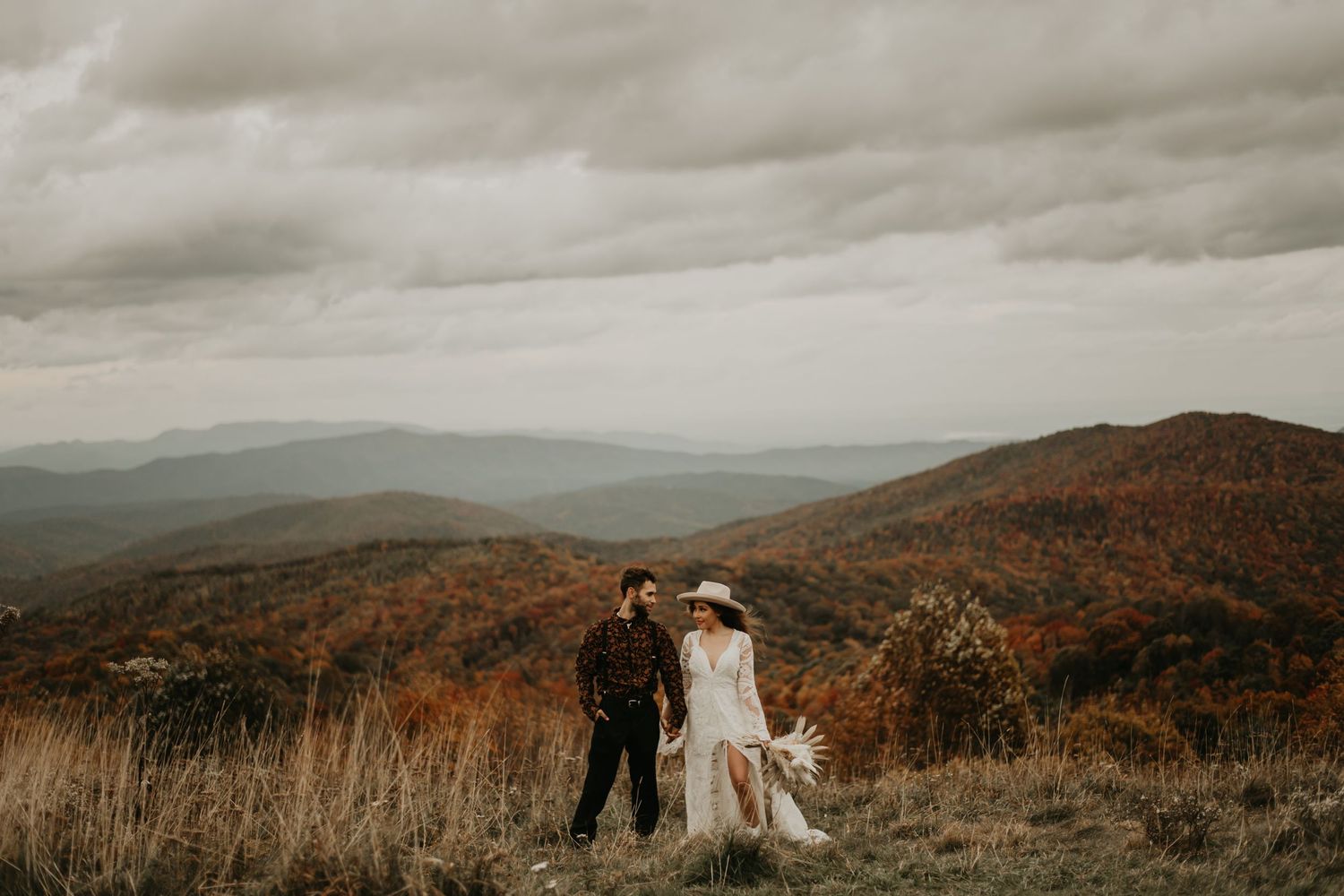 A couple in wedding attire stands together on a mountain overlook with rolling hills in the background in North Carolina.
