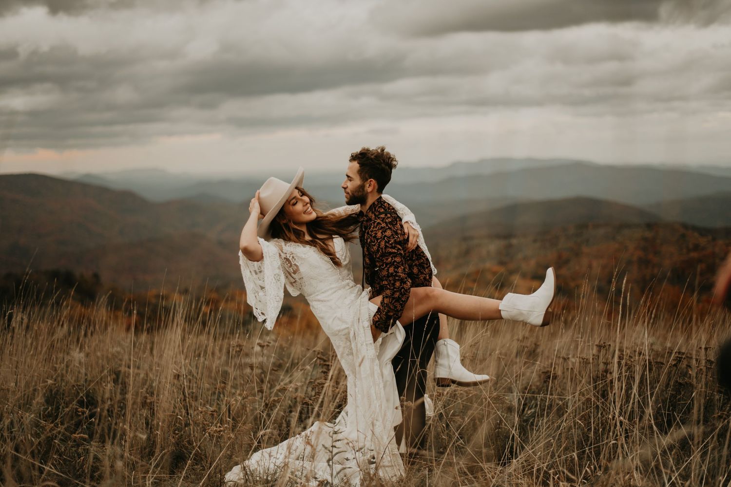 A playful wedding couple dances on a mountain ridge with dramatic autumn views in the background in North Carolina.