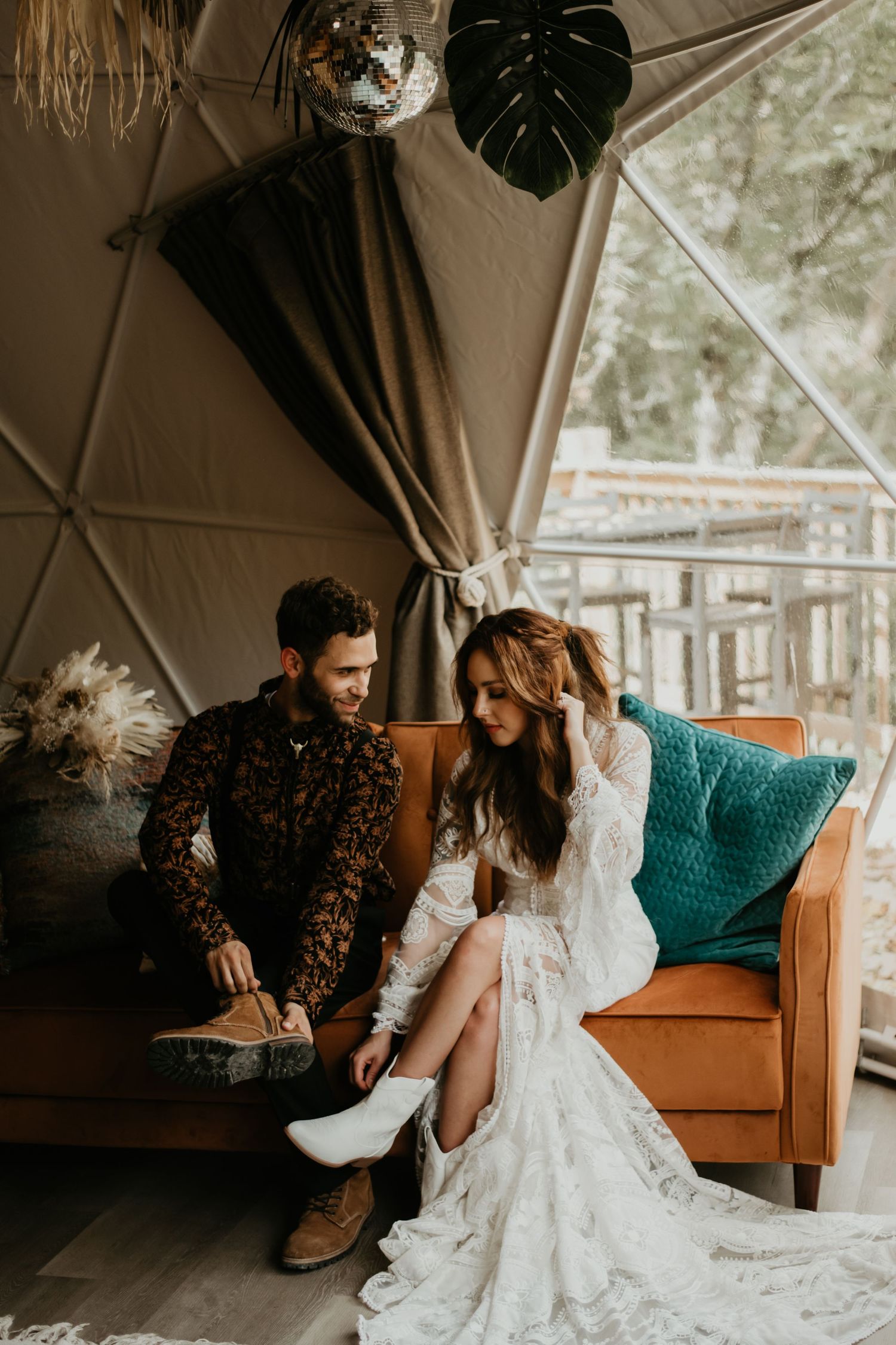 Wedding couple in boho attire sitting on leather couch under hanging monstera leaf in glamping tent.