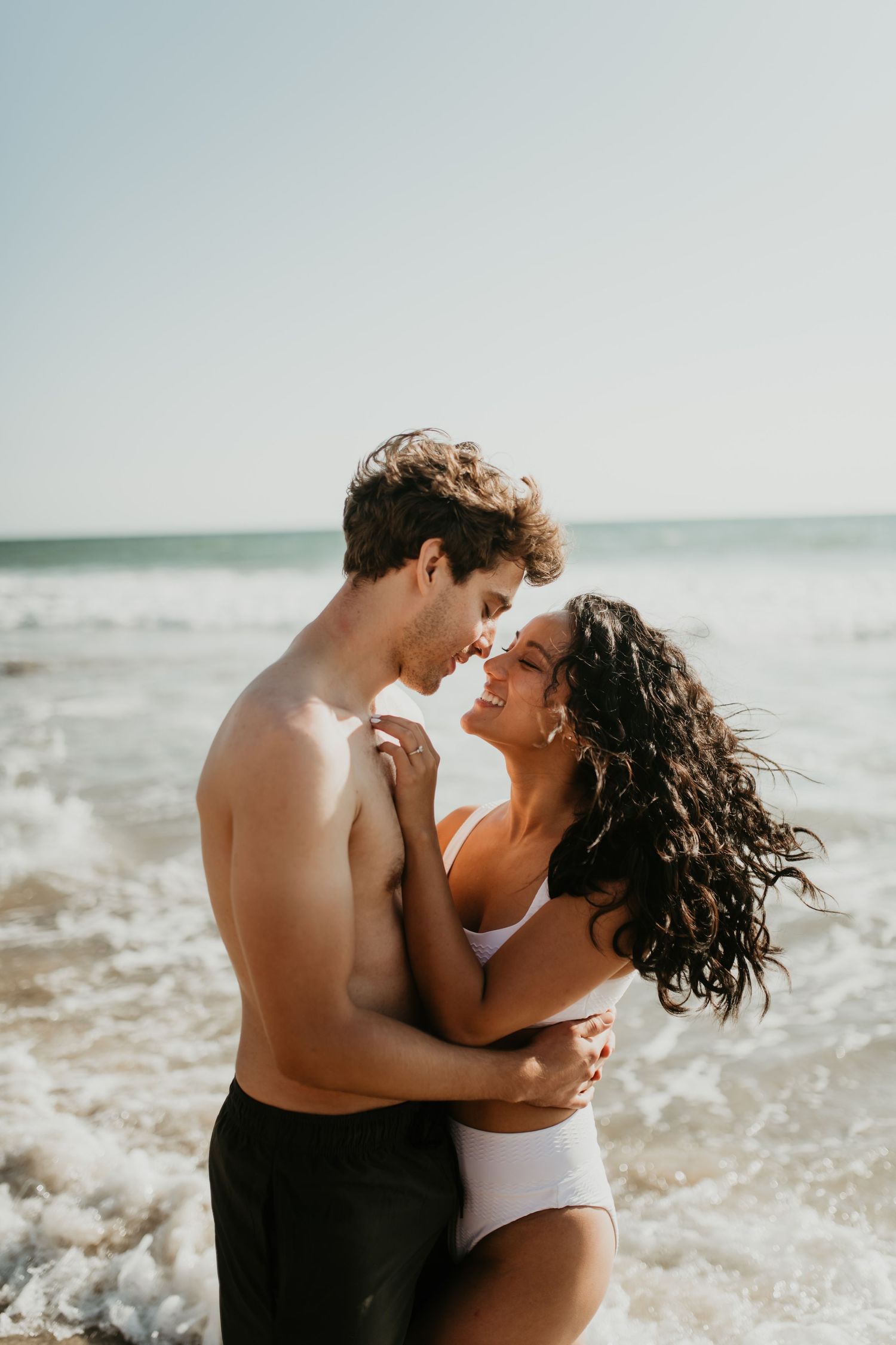 A romantic couple embraces on a sunny beach with ocean waves in the background.