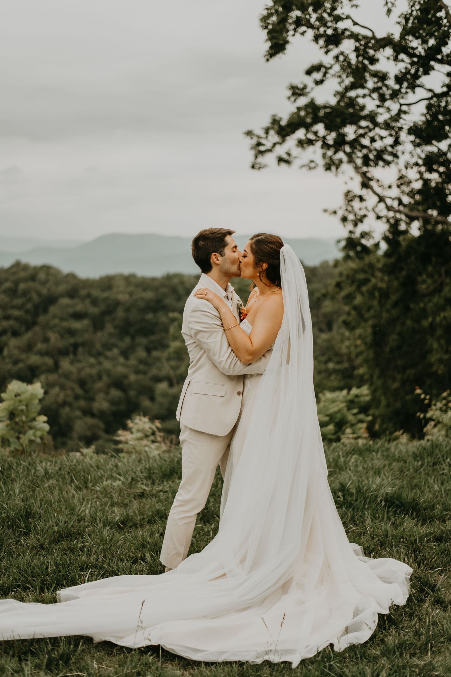 Couple in light wedding attire shares tender moment on hilltop with mountain backdrop.