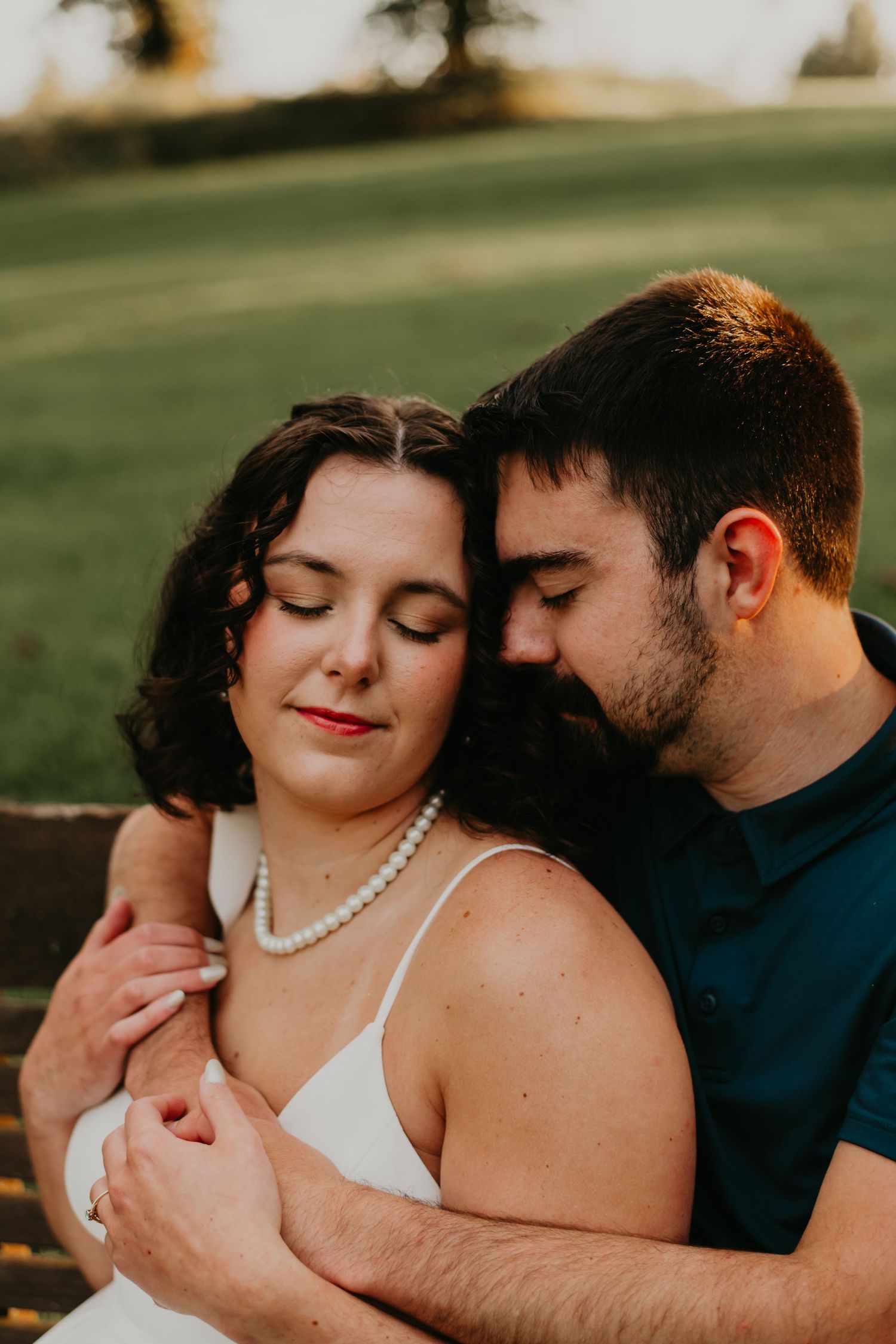 A romantic couple in wedding attire embraces tenderly in an outdoor setting with lush green backdrop.