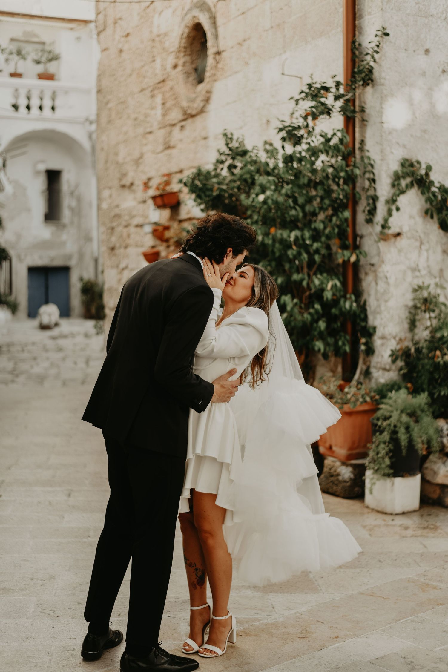 A romantic wedding kiss against an old Mediterranean stone wall with climbing plants in Italy.
