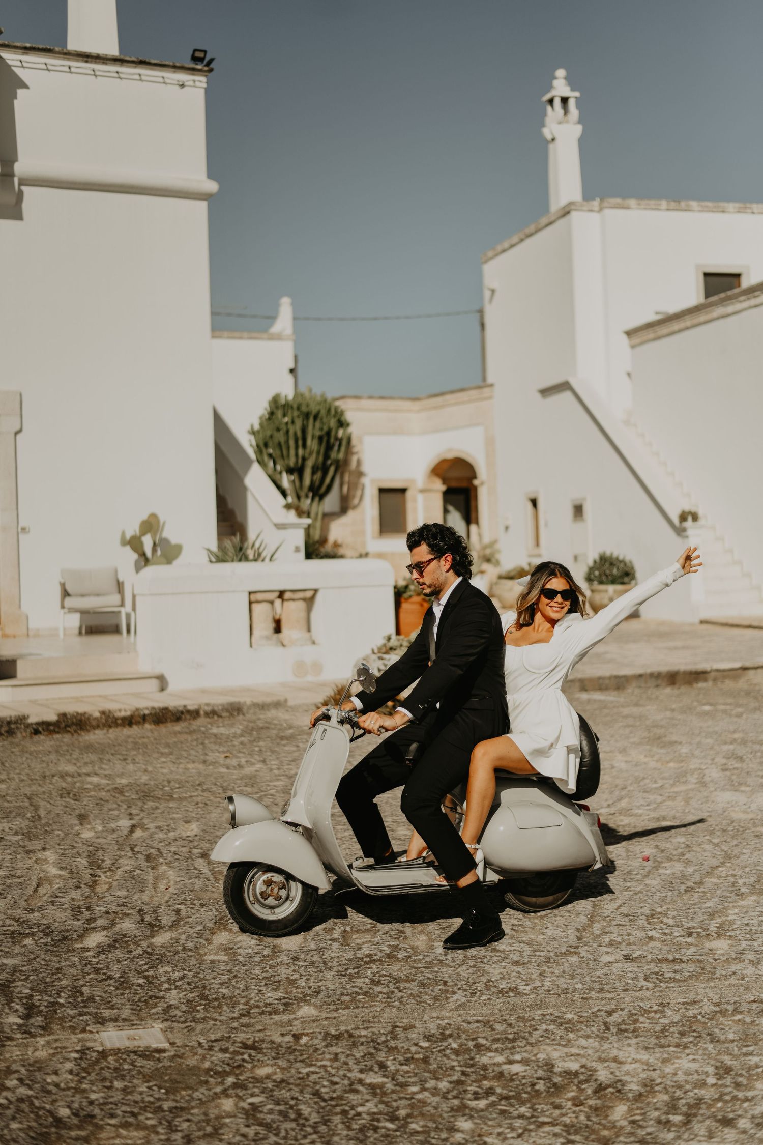 Couple riding white Vespa scooter through Mediterranean village with white buildings in Italy.