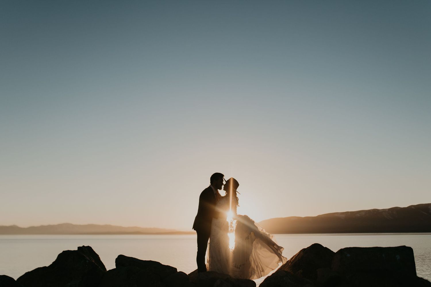 Silhouette of wedding couple embracing at sunset by rocky coastline.
