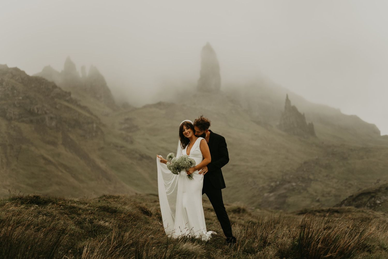 A wedding couple embraces on misty cliffs in dramatic mountainous landscape in Isle of Skye Scotland.