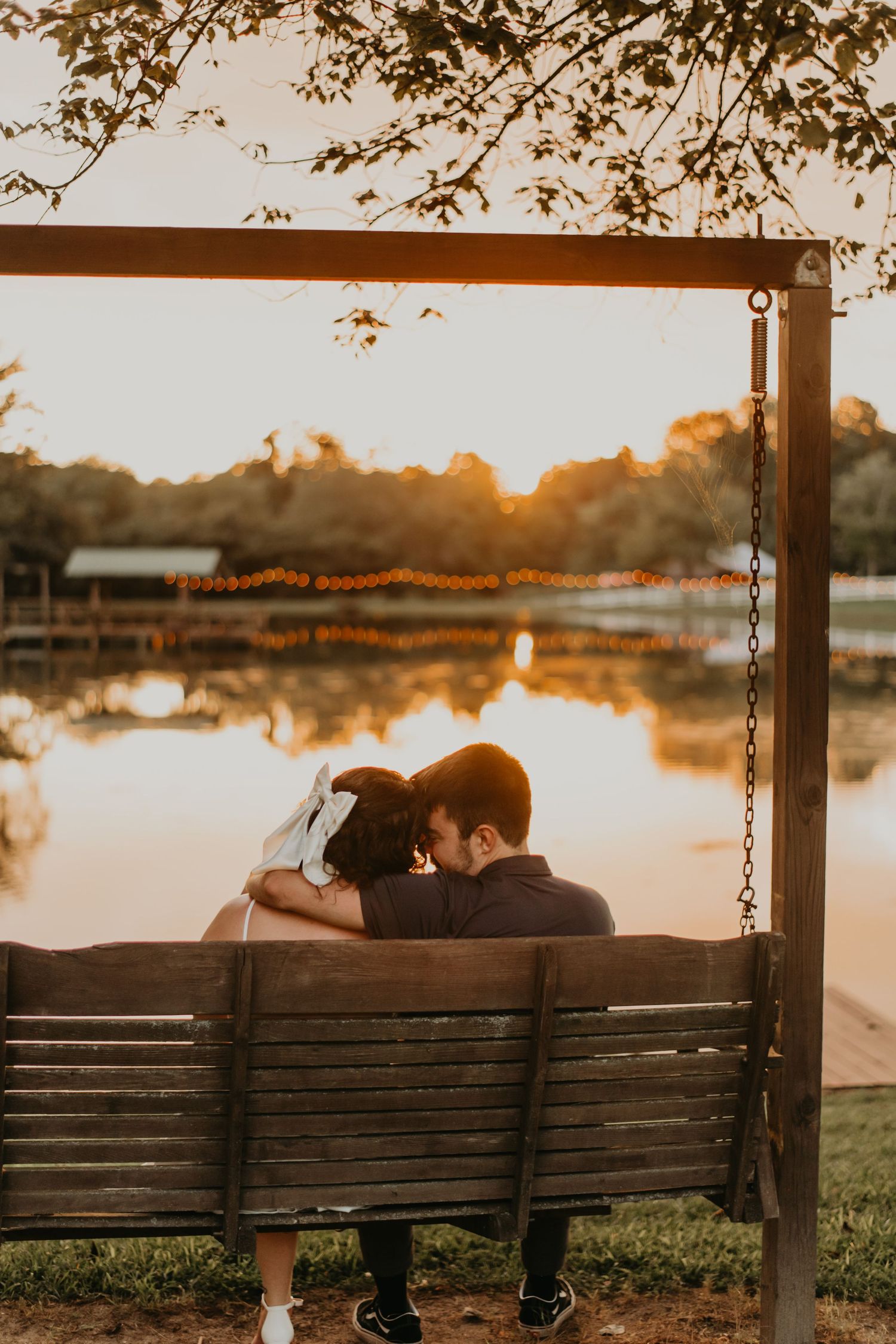 A couple sits together on a wooden porch swing overlooking a serene lake at sunset.