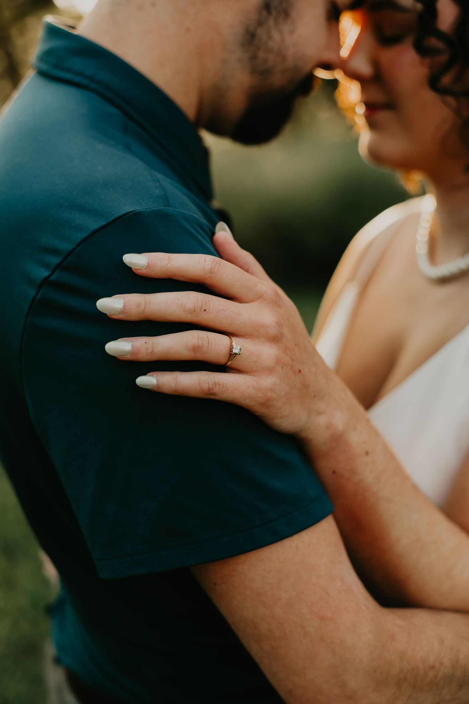 Close-up of a couple's intimate moment showing hands with wedding ring against a teal shirt in warm sunlight.