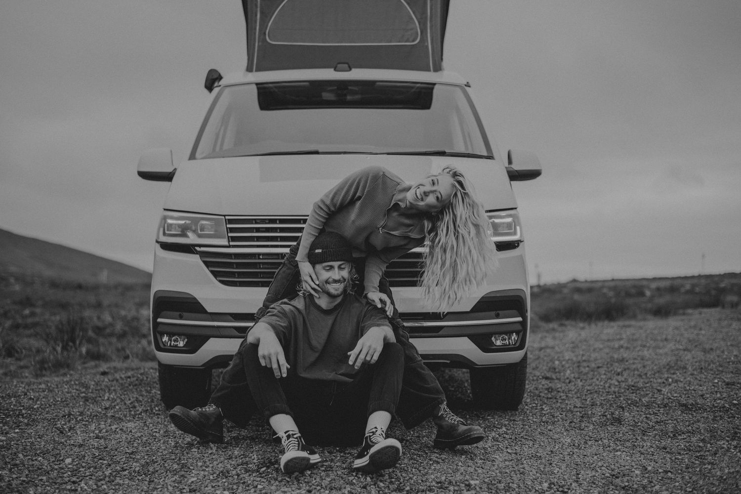 Black and white photo of someone crouching in front of a parked van on a hillside in moody weather.