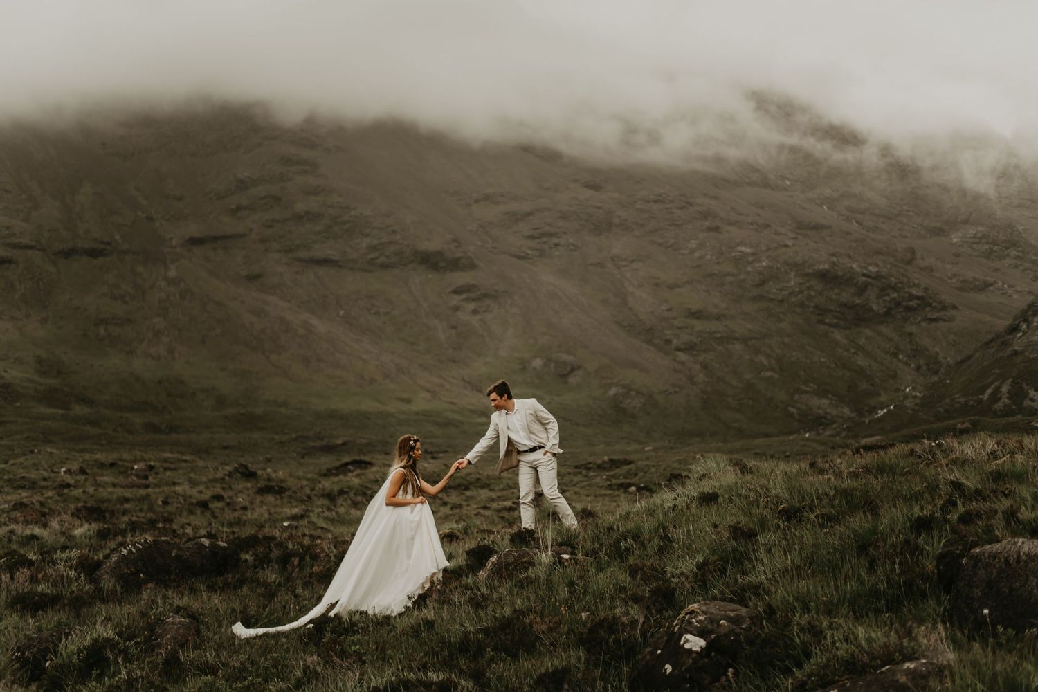 A couple in wedding attire walking hand in hand through misty mountain terrain in isle of Skye Scotland.