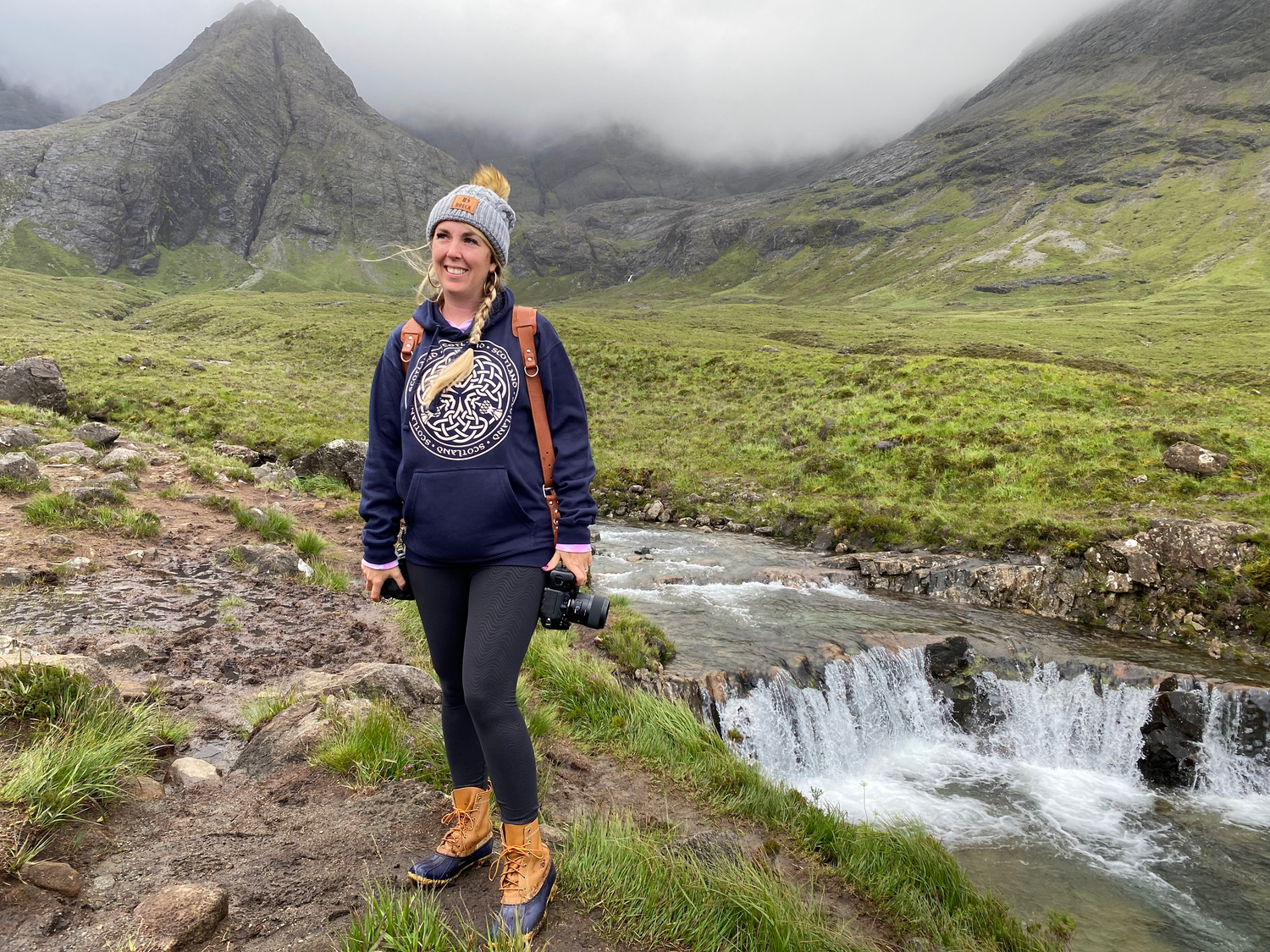 A hiker stands smiling by a waterfall in the Scottish Highlands with mountains in the background in isle of Skye Scotland.