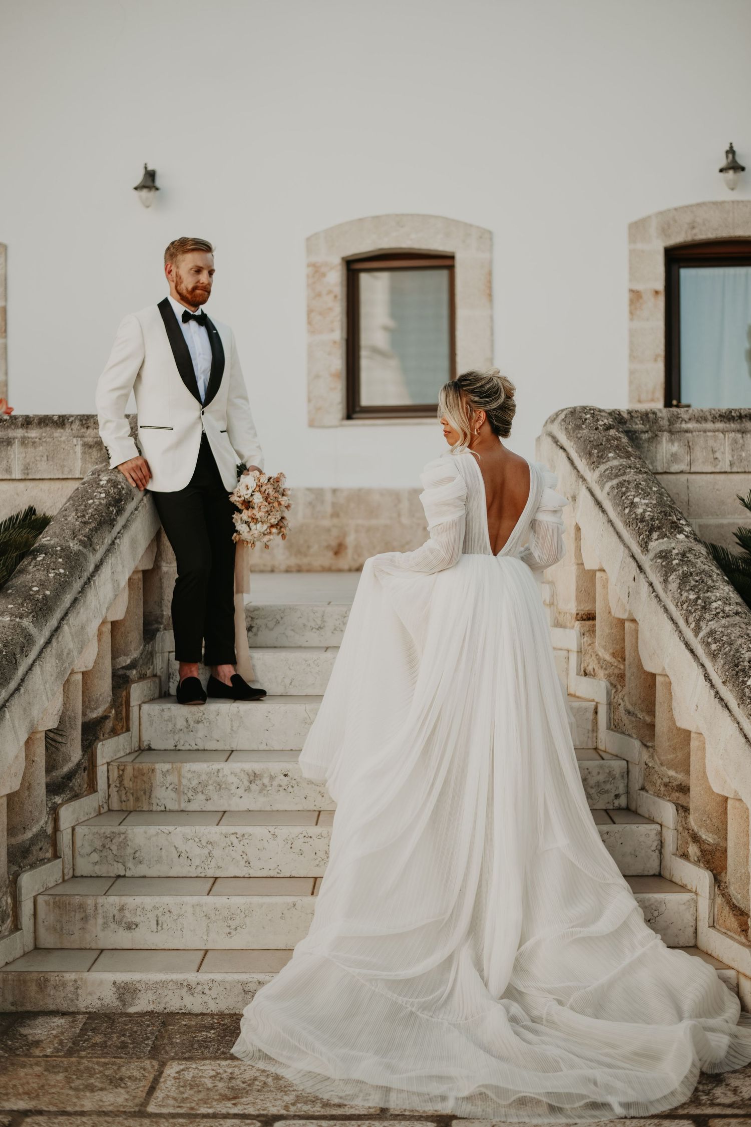 A wedding couple poses on elegant stone stairs with the bride's flowing white dress in focus in Puglia Italy.