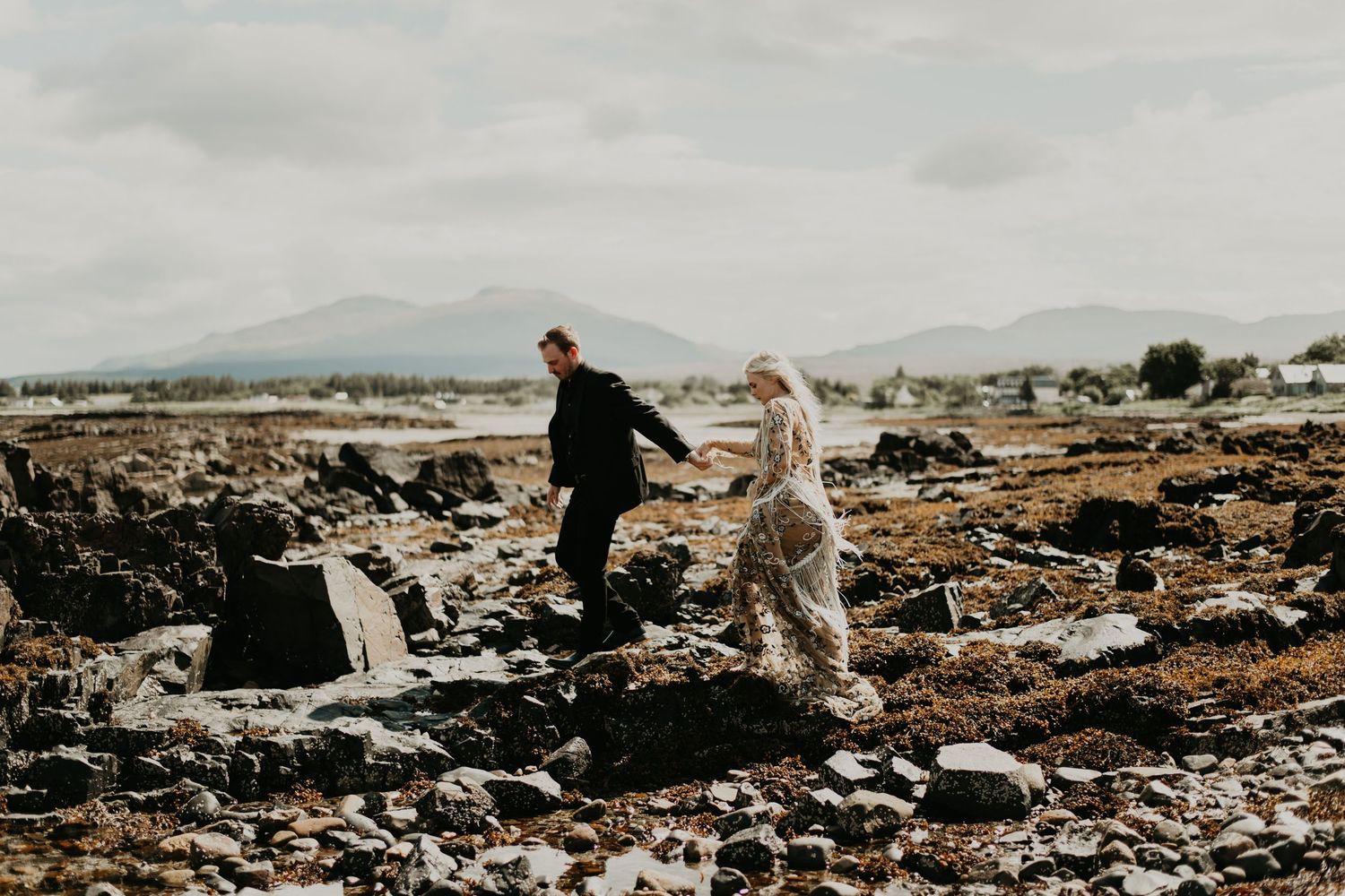 A couple explores rocky terrain together on their wedding day with mountains in the distance in isle of Skye Scotland.