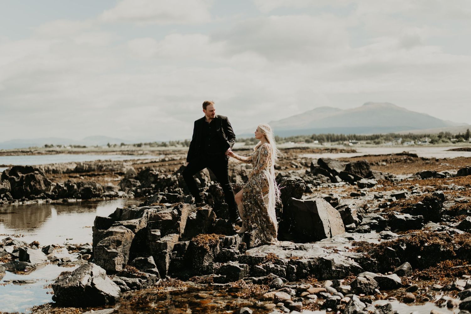 A couple stands on dramatic rocky coastal tidepools with mountains in the distance in Isle of Skye Scotland.