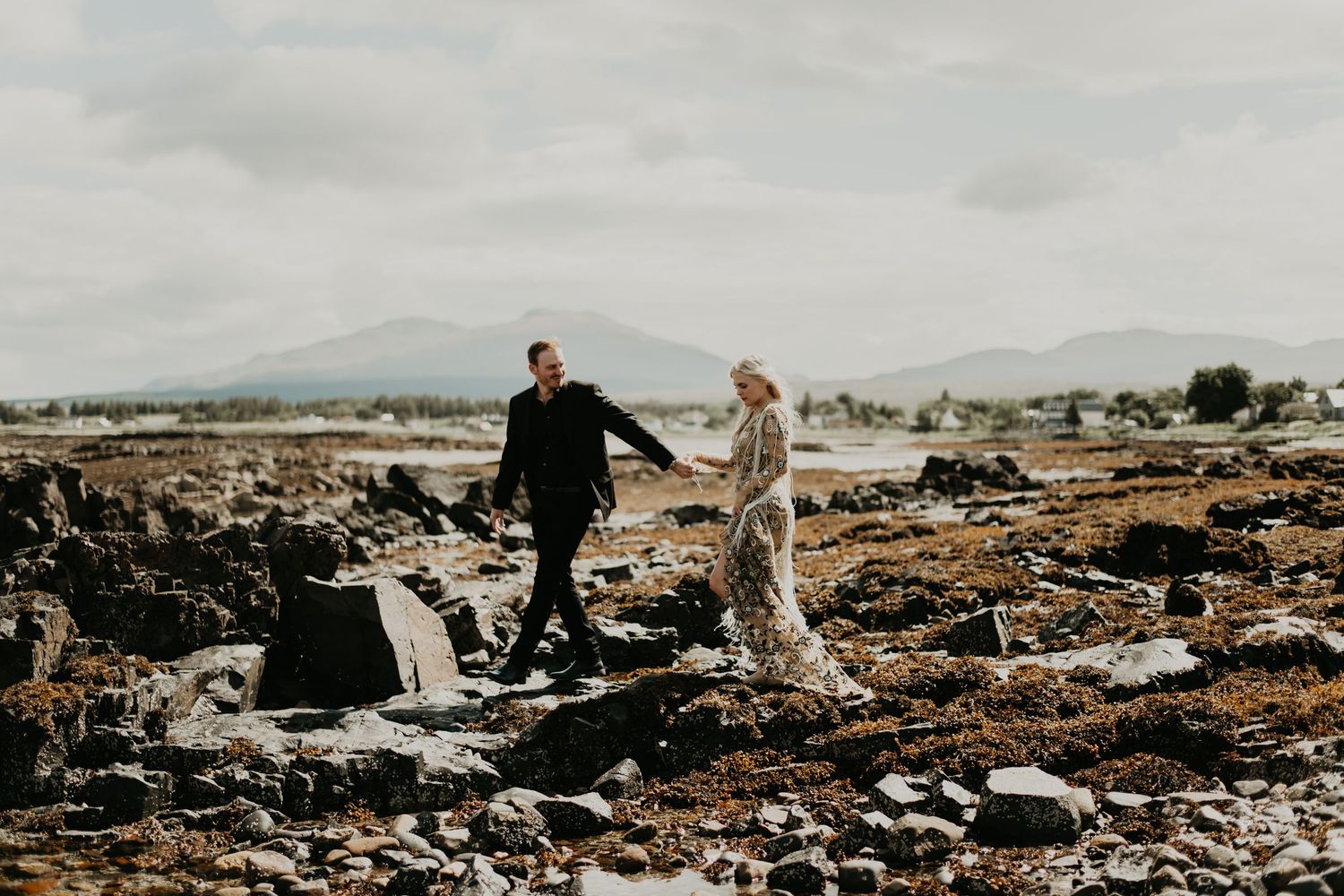 A couple walks hand in hand across rocky terrain with mountains in the background in isle of skye Scotland.