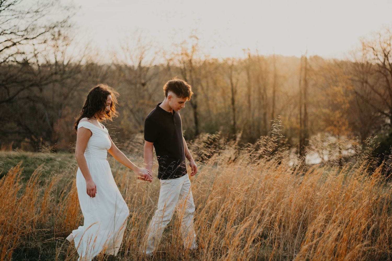 Two people walk through a golden field of tall grass at sunset with warm autumn light glowing in the background.