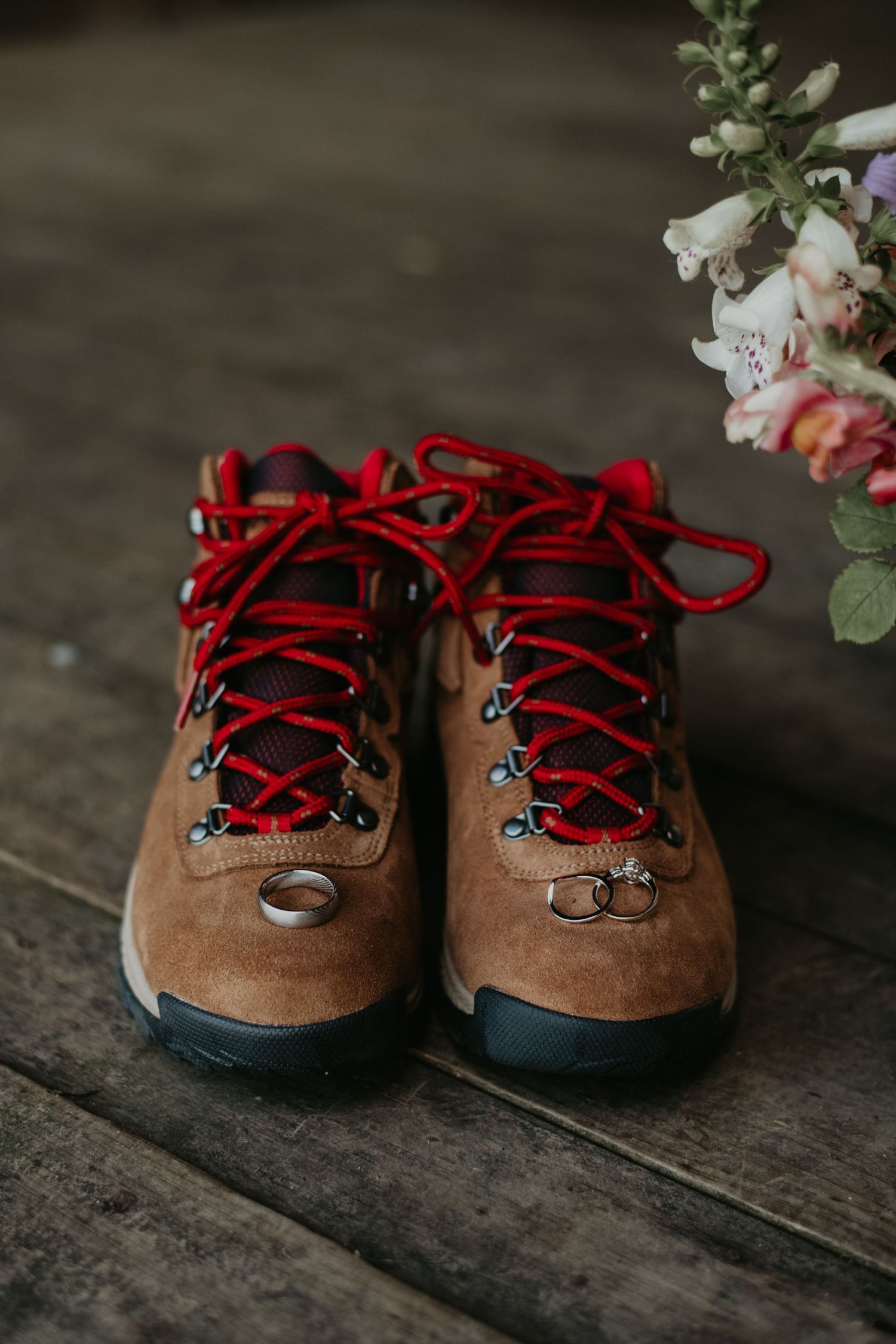 Brown suede hiking boots with bright red laces on wooden floor with soft floral arrangement in background.
