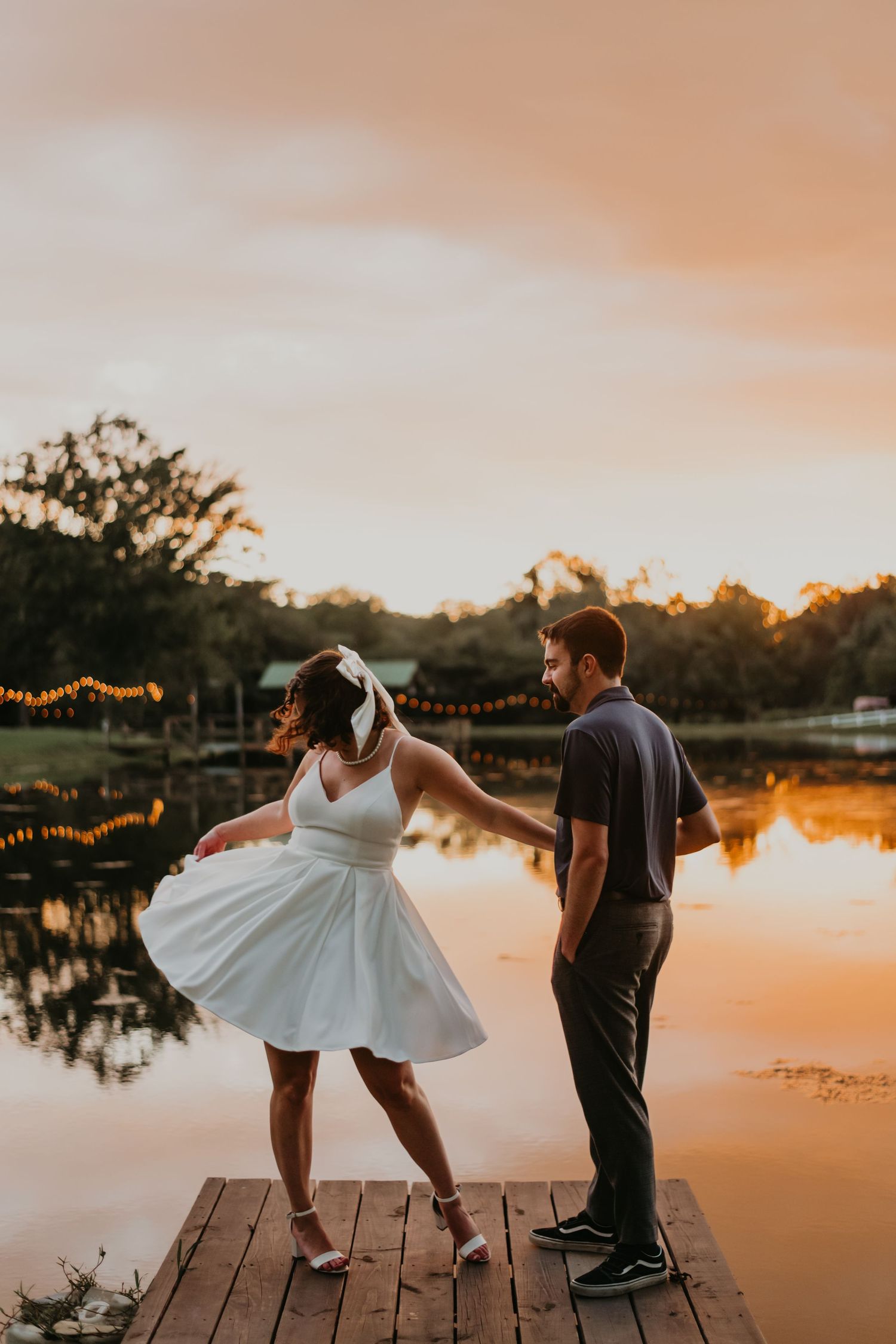 Couple dancing and posing on wooden dock at sunset with string lights overhead creating a romantic atmosphere.