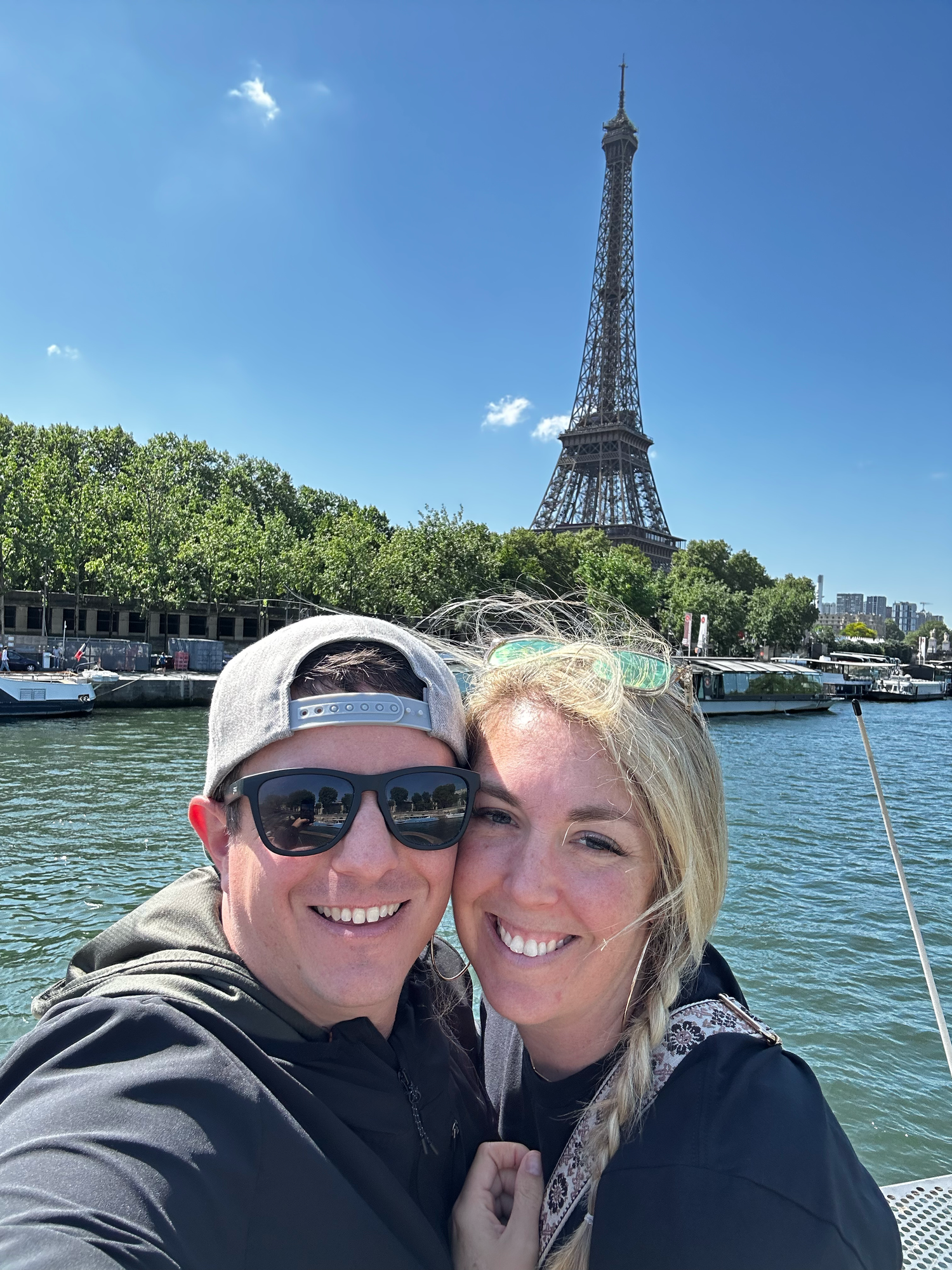 Couple taking selfie on boat with Eiffel Tower and Seine River in background.