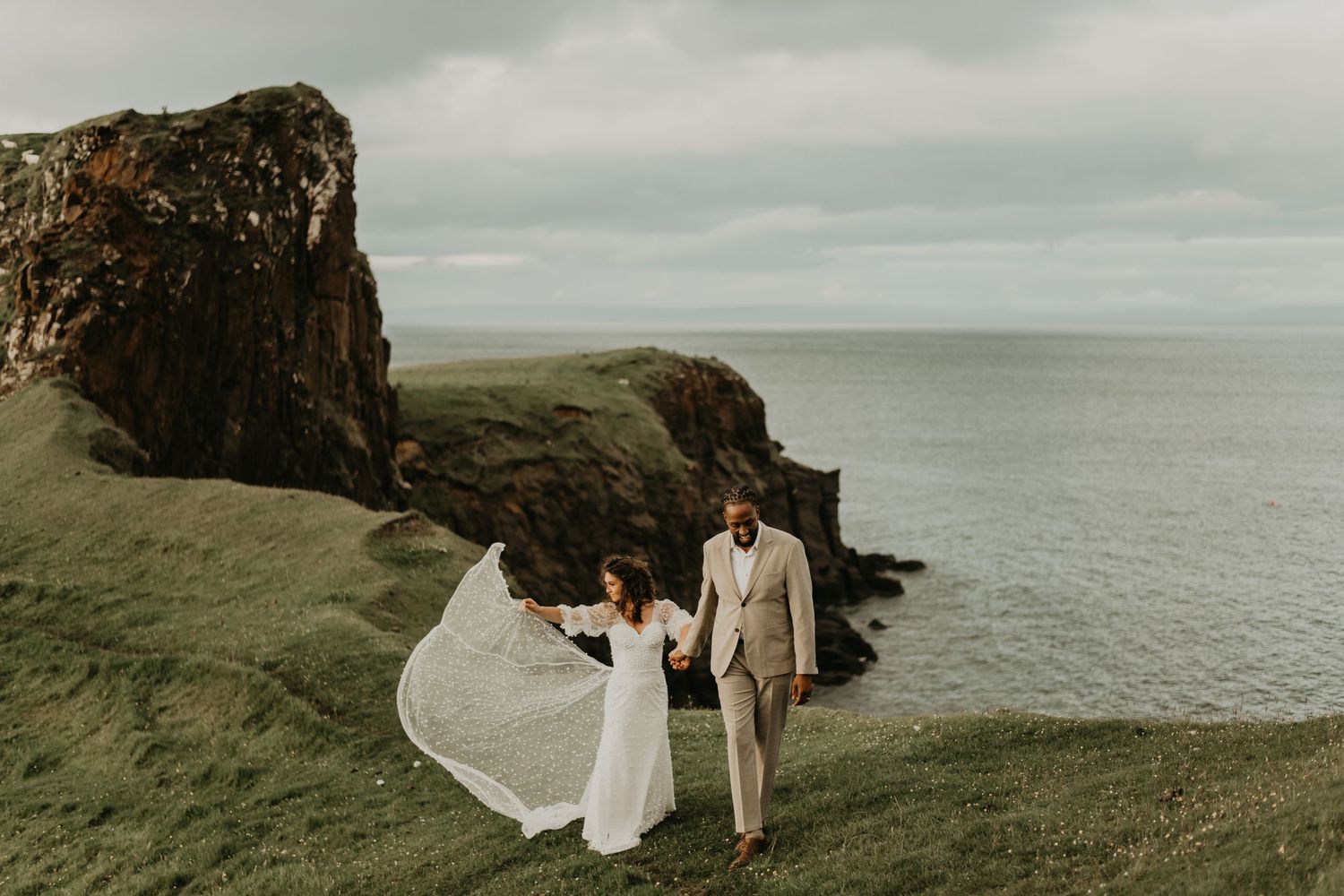 A white wedding dress flows in the wind as a couple stands on dramatic coastal cliffs overlooking the ocean in isle of Skye Scotland.