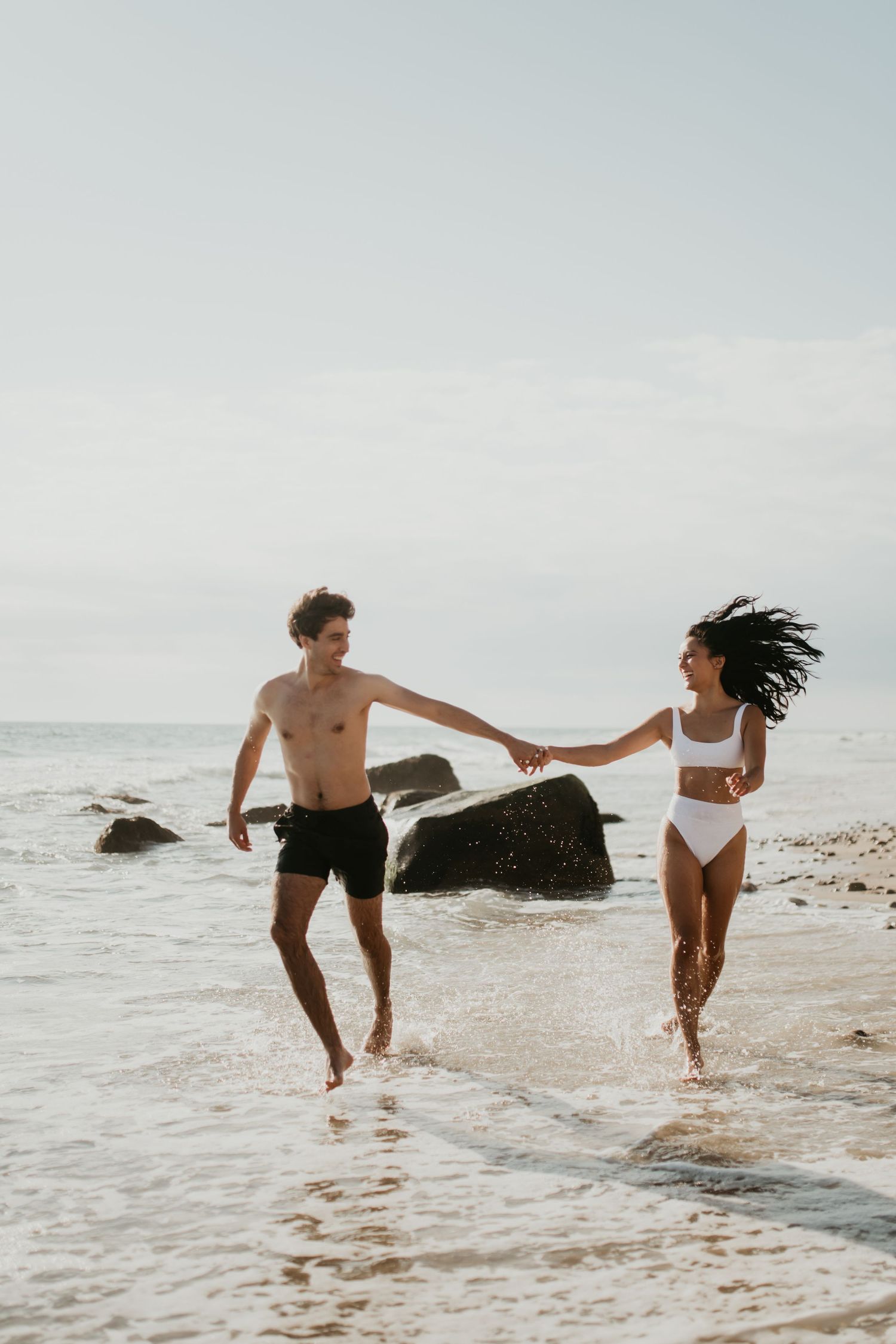 A couple in swimwear holds hands while running joyfully along a rocky beach during a cloudy day.