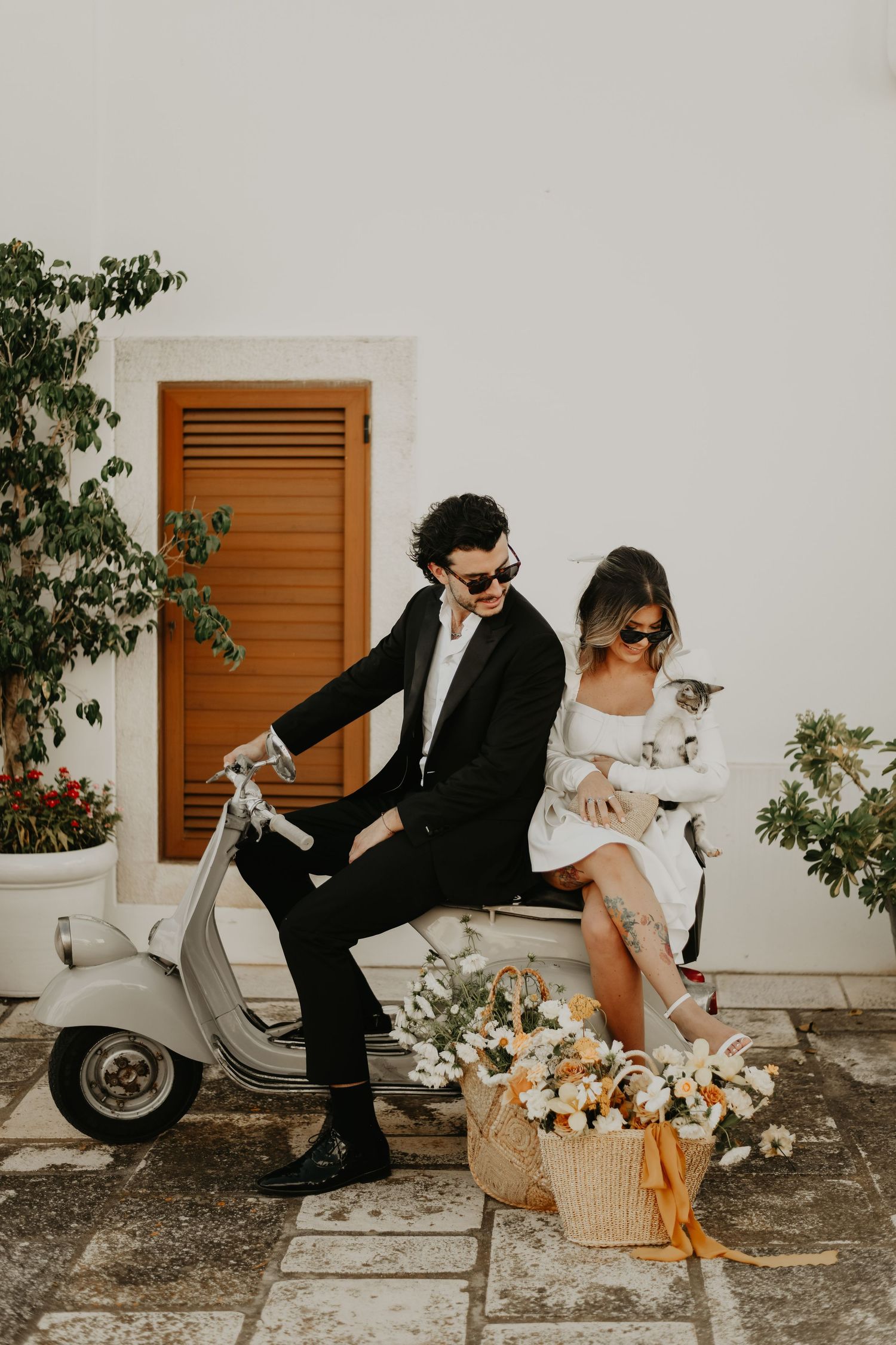 Stylish couple in sunglasses poses with a vintage Vespa scooter and flower basket in Italy.