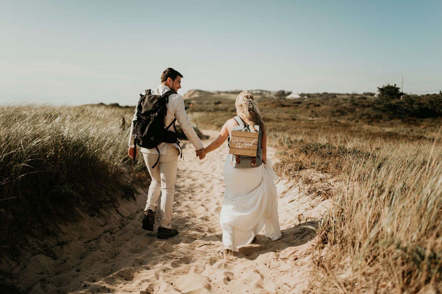 A couple walks through coastal sand dunes holding hands at sunset.