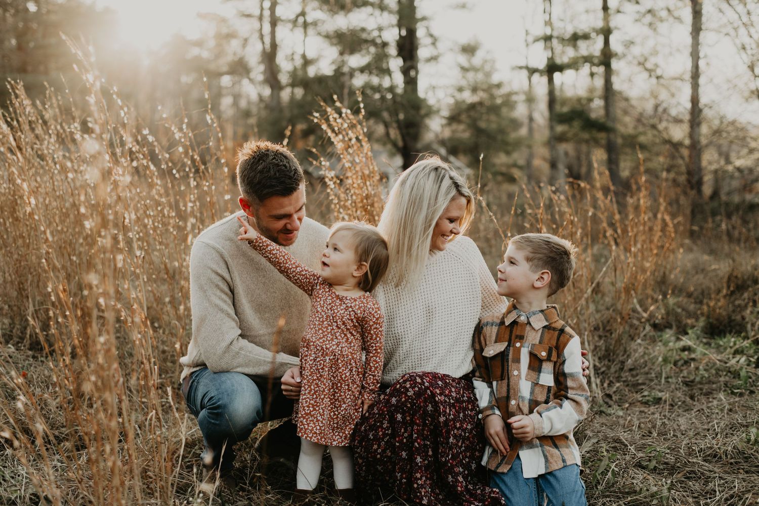 A loving family shares a tender moment together in a sunlit field with tall golden grass and evergreen trees.