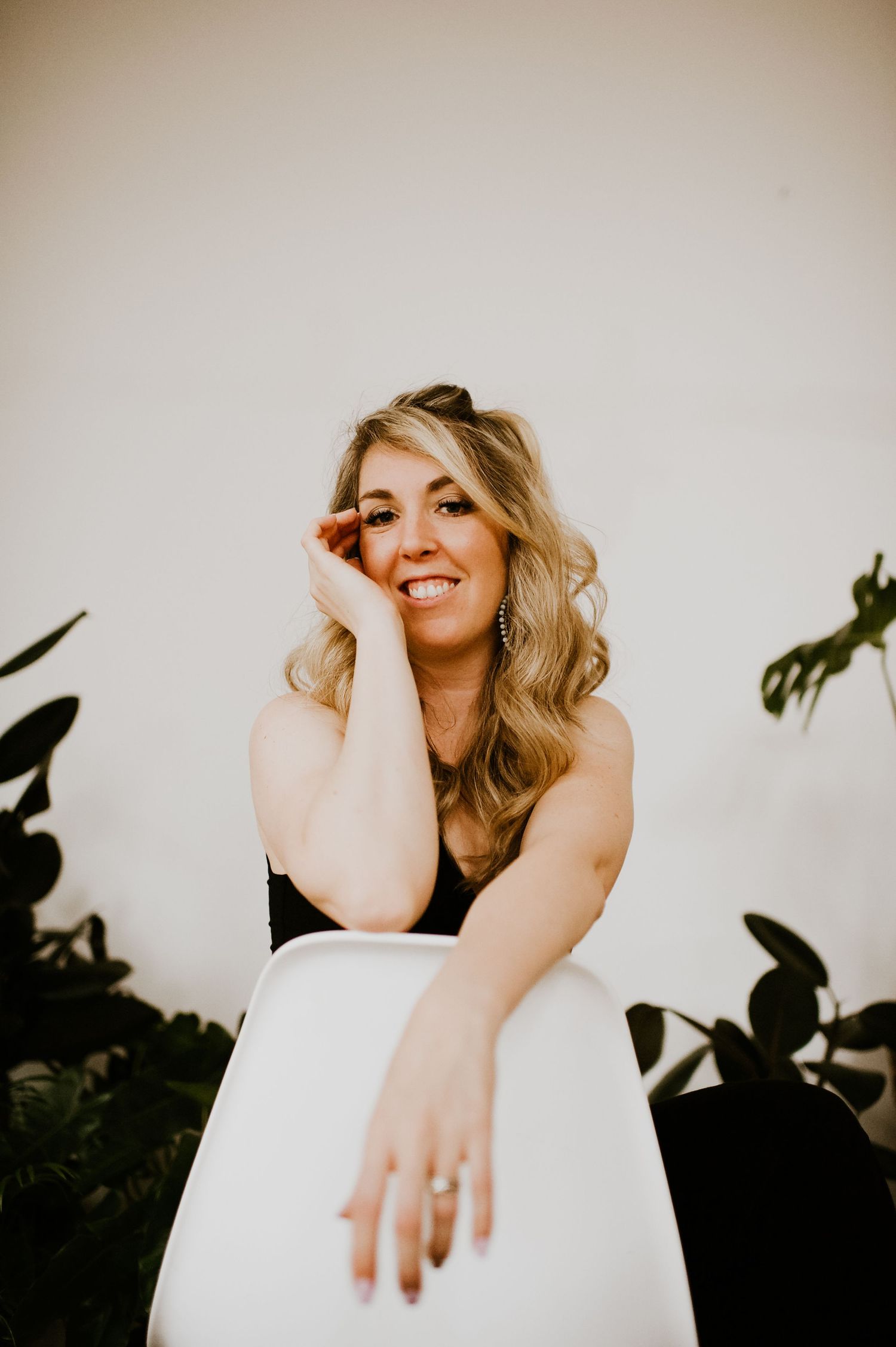 A person in a black and white dress poses cheerfully against a neutral wall with plants.
