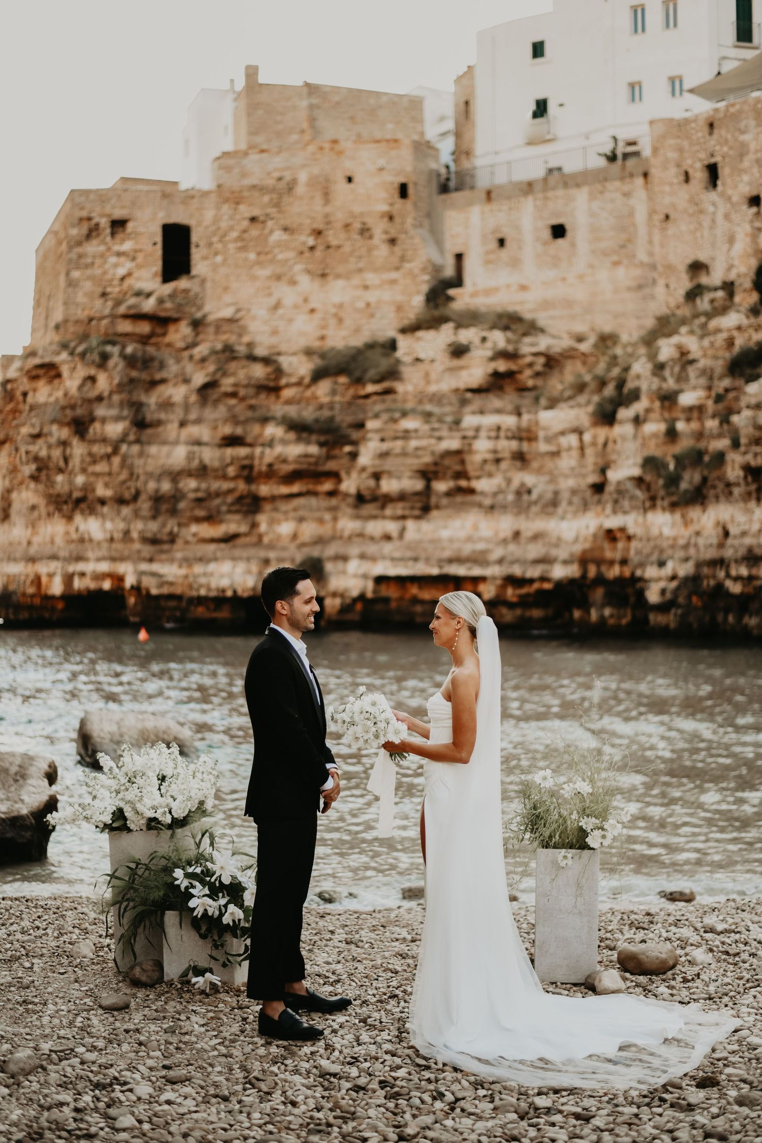 Wedding ceremony on rocky coastline with ancient stone buildings perched on cliffs above in Italy.