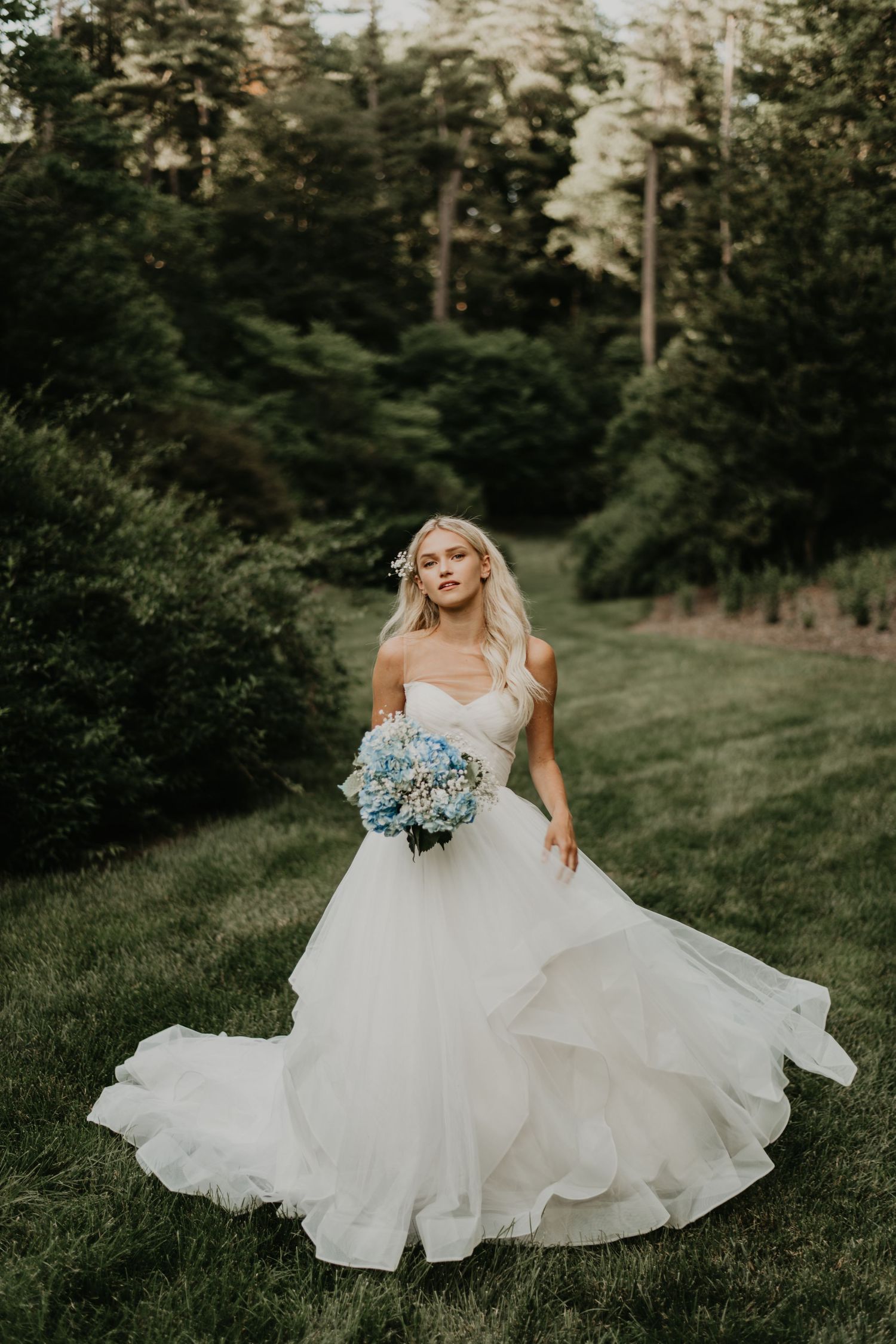 A bride in a flowing white ballgown poses with blue hydrangea bouquet against a wooded backdrop in Asheville north Carolina.