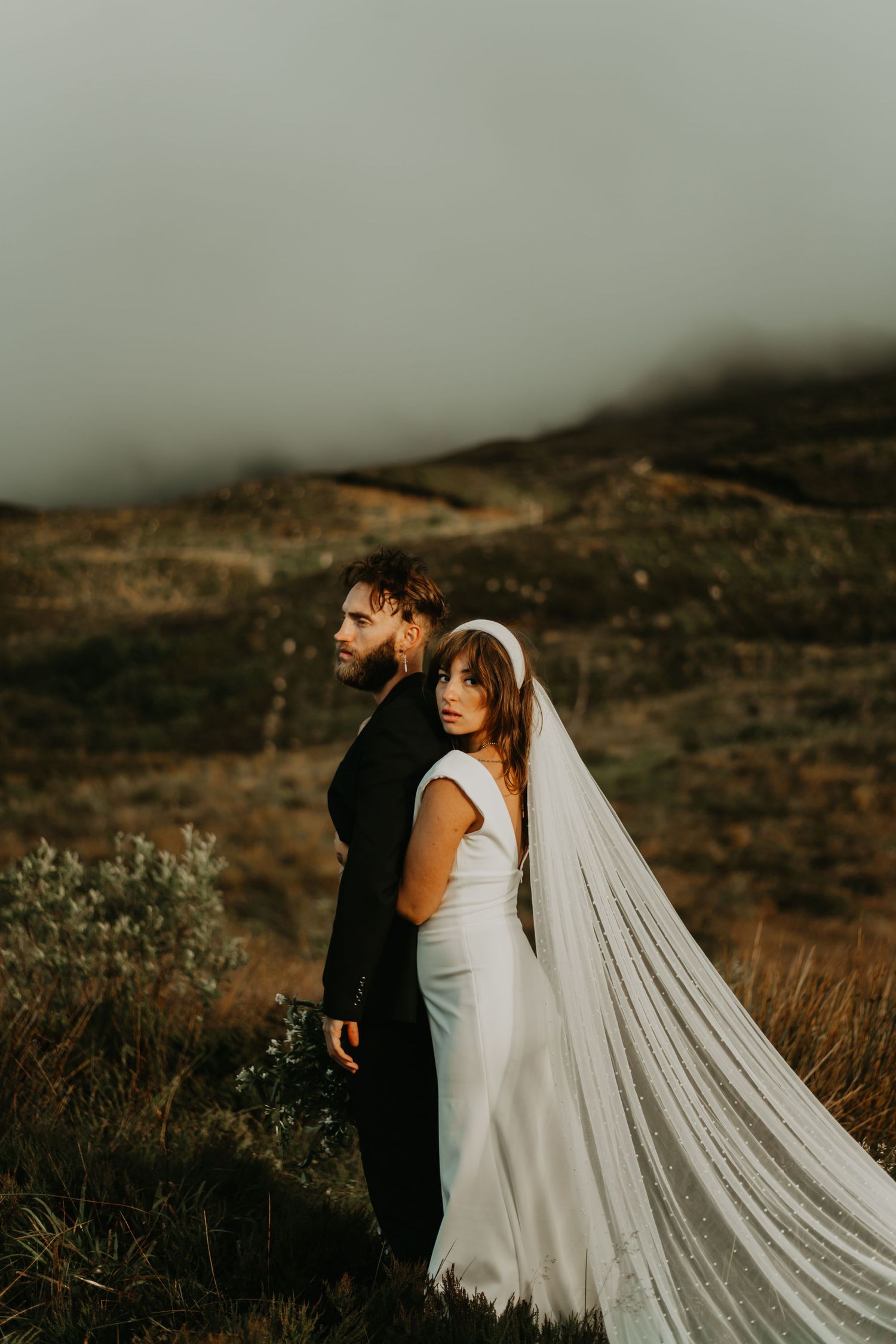 A couple in wedding attire embrace against a misty mountainous backdrop as a long white veil flows in the wind.