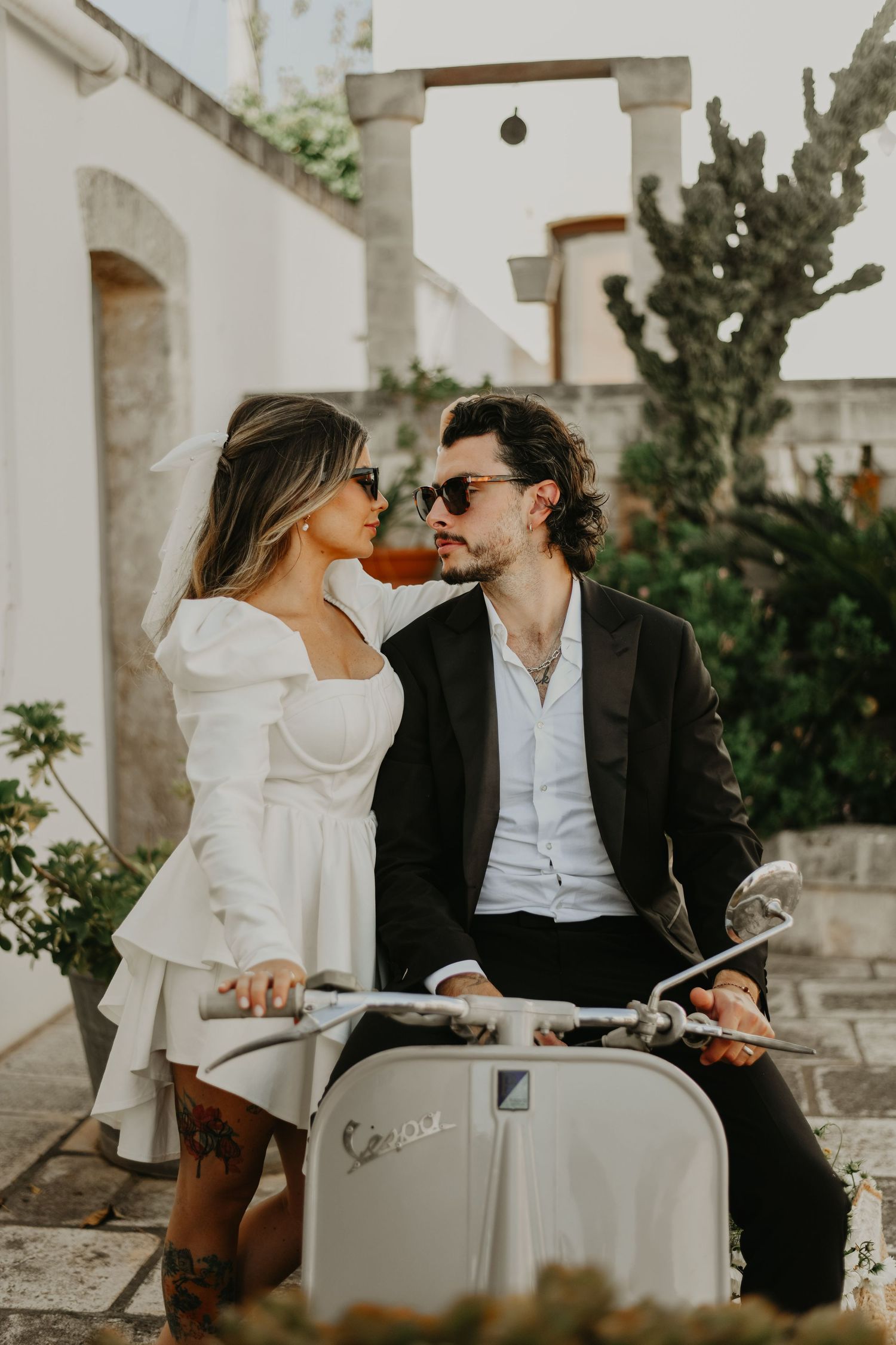 Stylish couple in formal wear sitting intimately on vintage Vespa scooter in Italy.