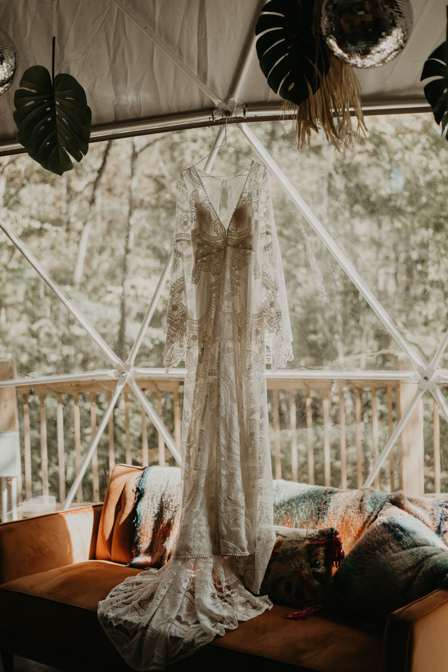 Delicate lace wedding dress hanging in a tent with tropical leaves and golden hour lighting.