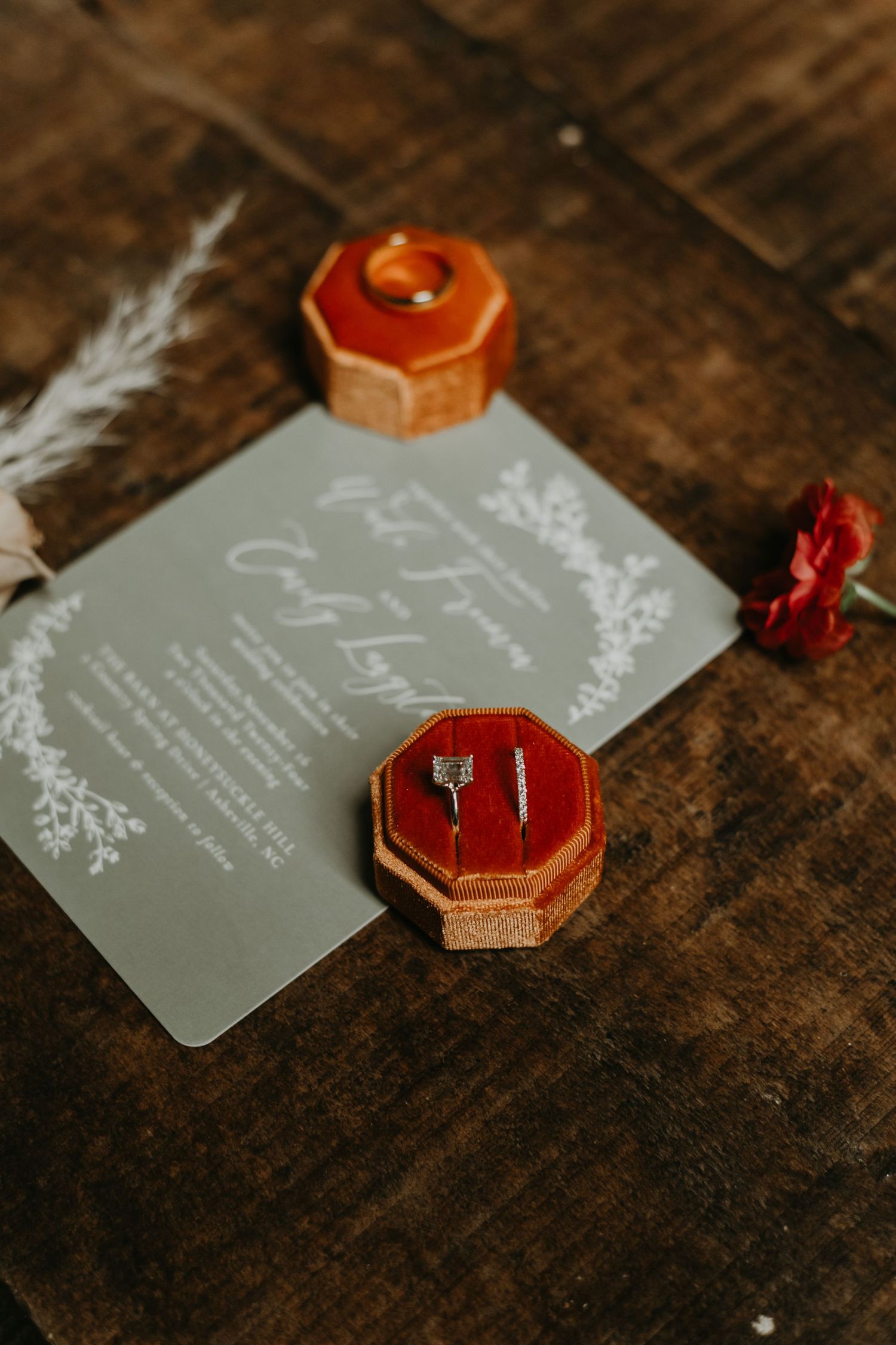Wedding ring displayed in orange velvet ring box next to gray invitation card on rustic wooden surface.